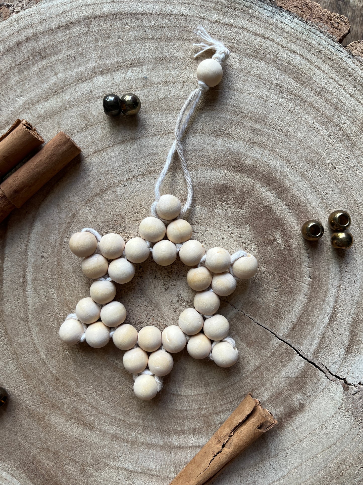 Small star-shaped decorative item made of small wooden beads and cotton yarn, on a wooden surface with cinnamon sticks and gold beads for decoration.
