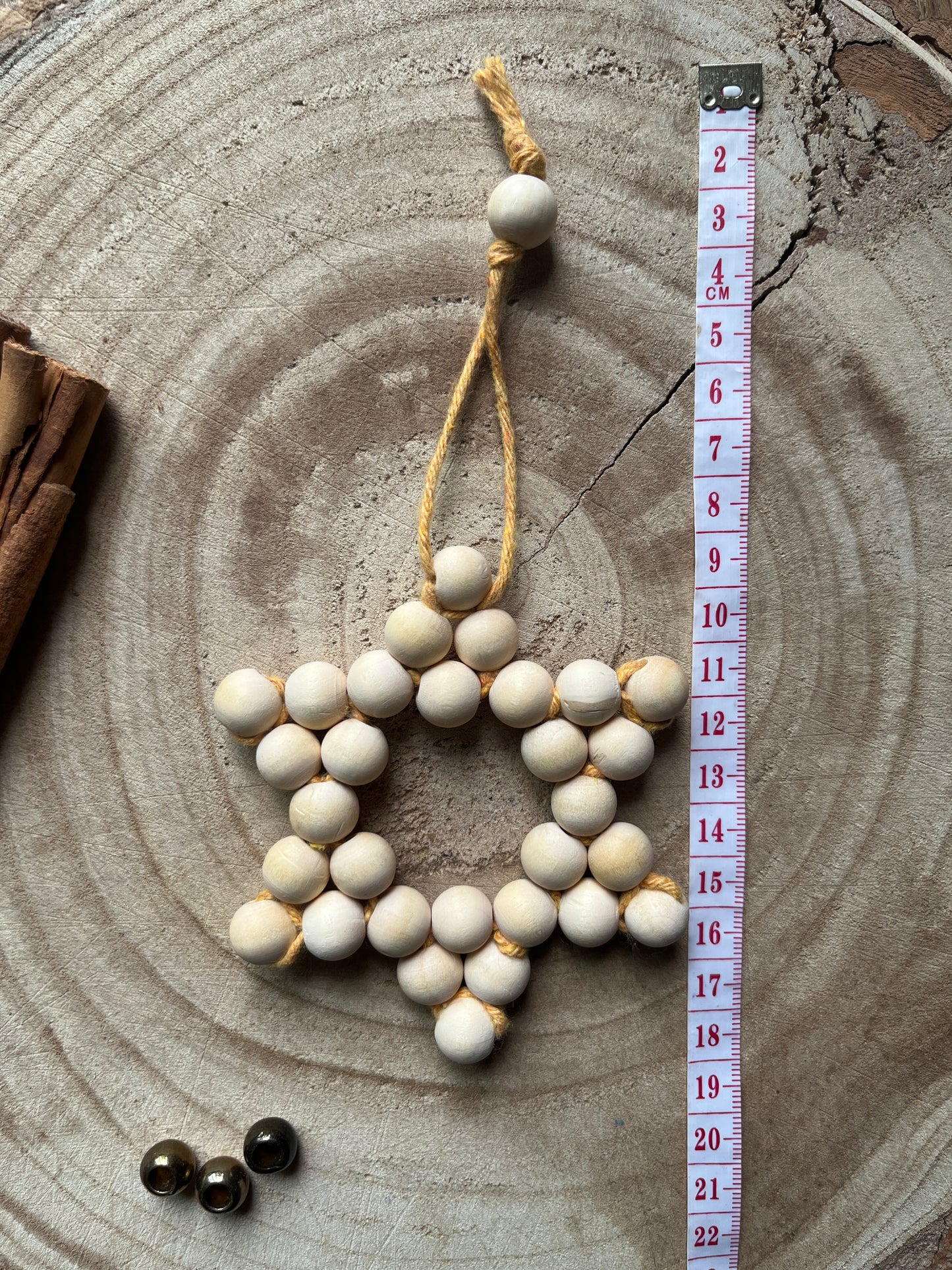 Star-shaped decorative item made of small wooden beads and yellow yarn, on a wooden surface with cinnamon sticks and gold beads for decoration, and a tape measure for size reference.