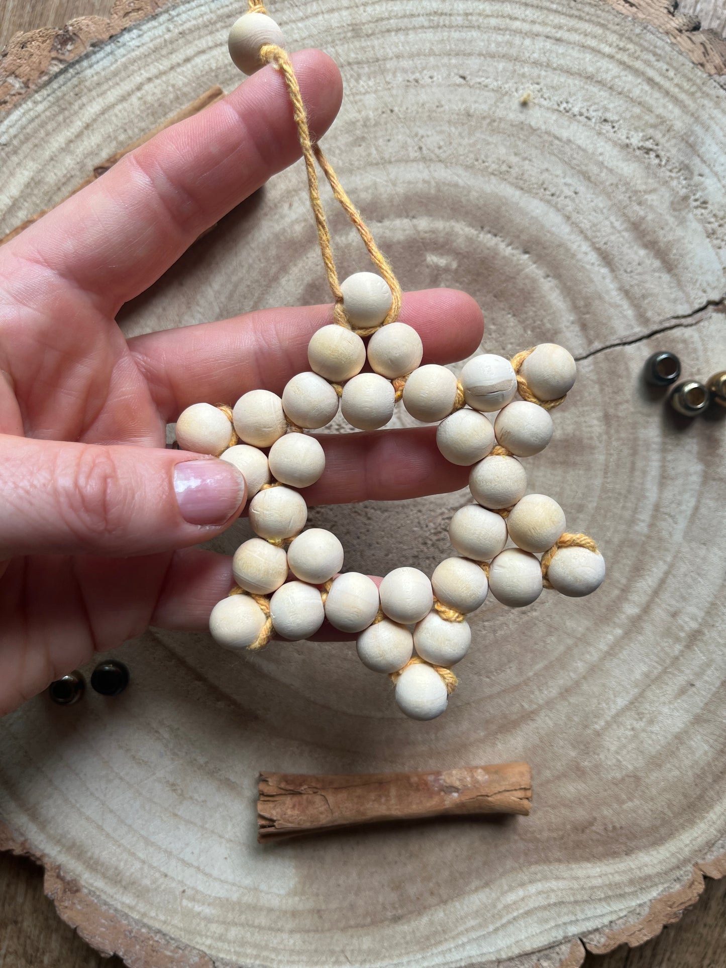 A hand holding a star-shaped decorative item made of small wooden beads and yellow yarn, on a wooden surface with cinnamon sticks and gold beads for decoration.