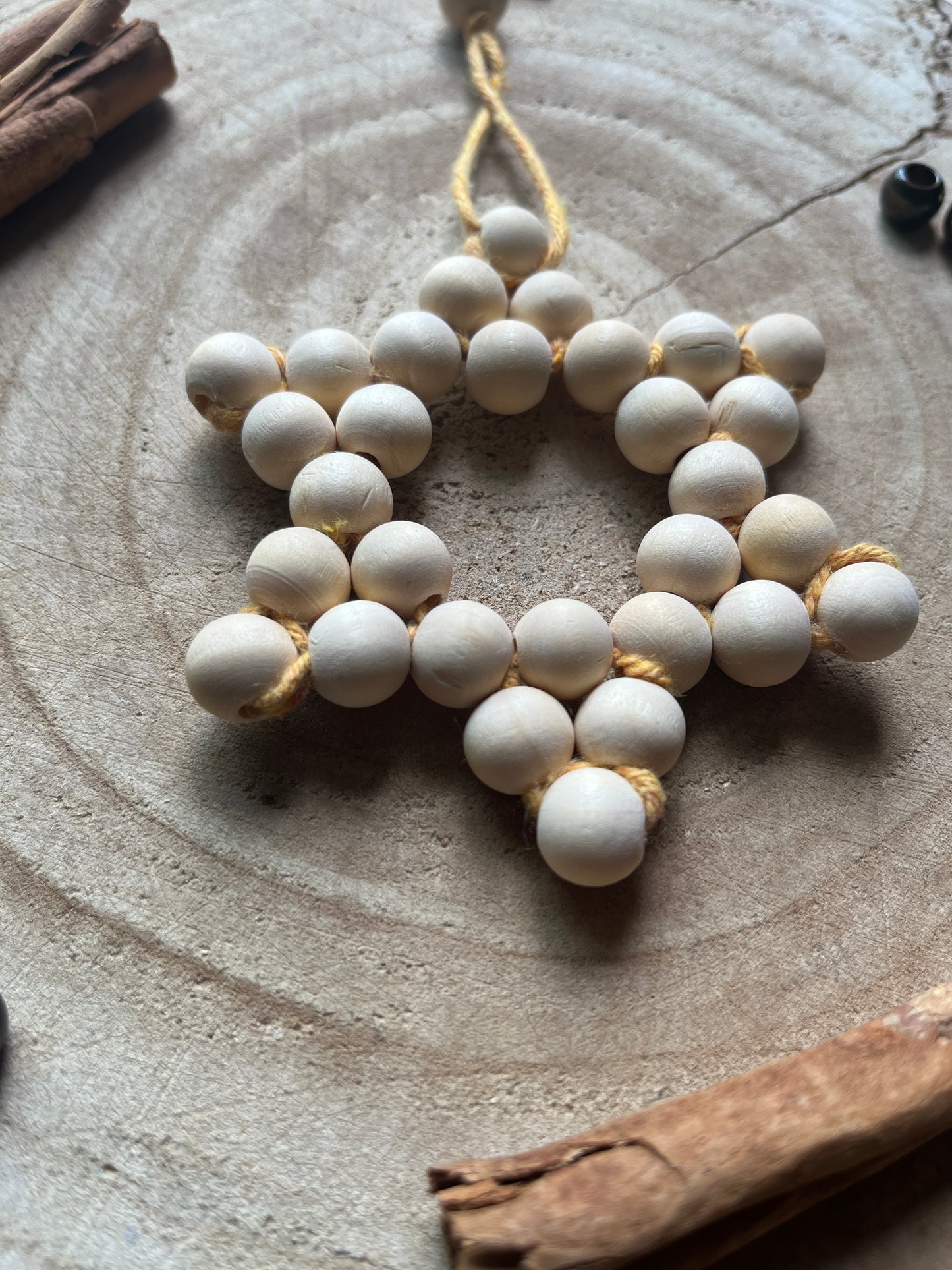 Close up of a star-shaped decorative item made of small wooden beads and yellow yarn, on a wooden surface with cinnamon sticks and gold beads for decoration.
