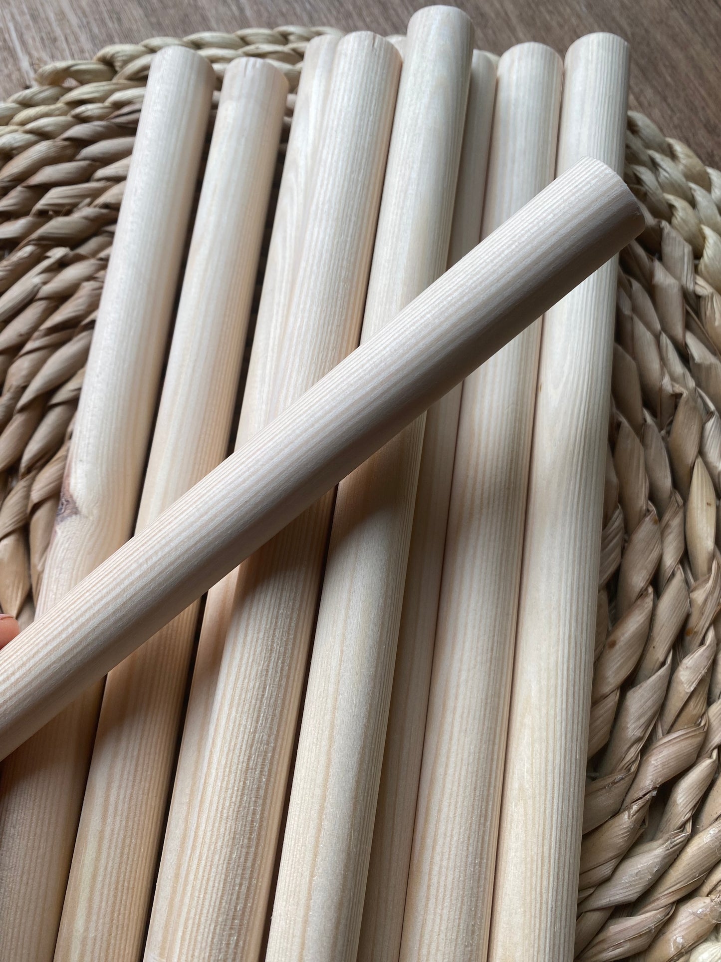 A pile of natural pine wooden dowels for macramé and crafts laid across a round wicker mat. One dowel lays across the others in the opposite direction.