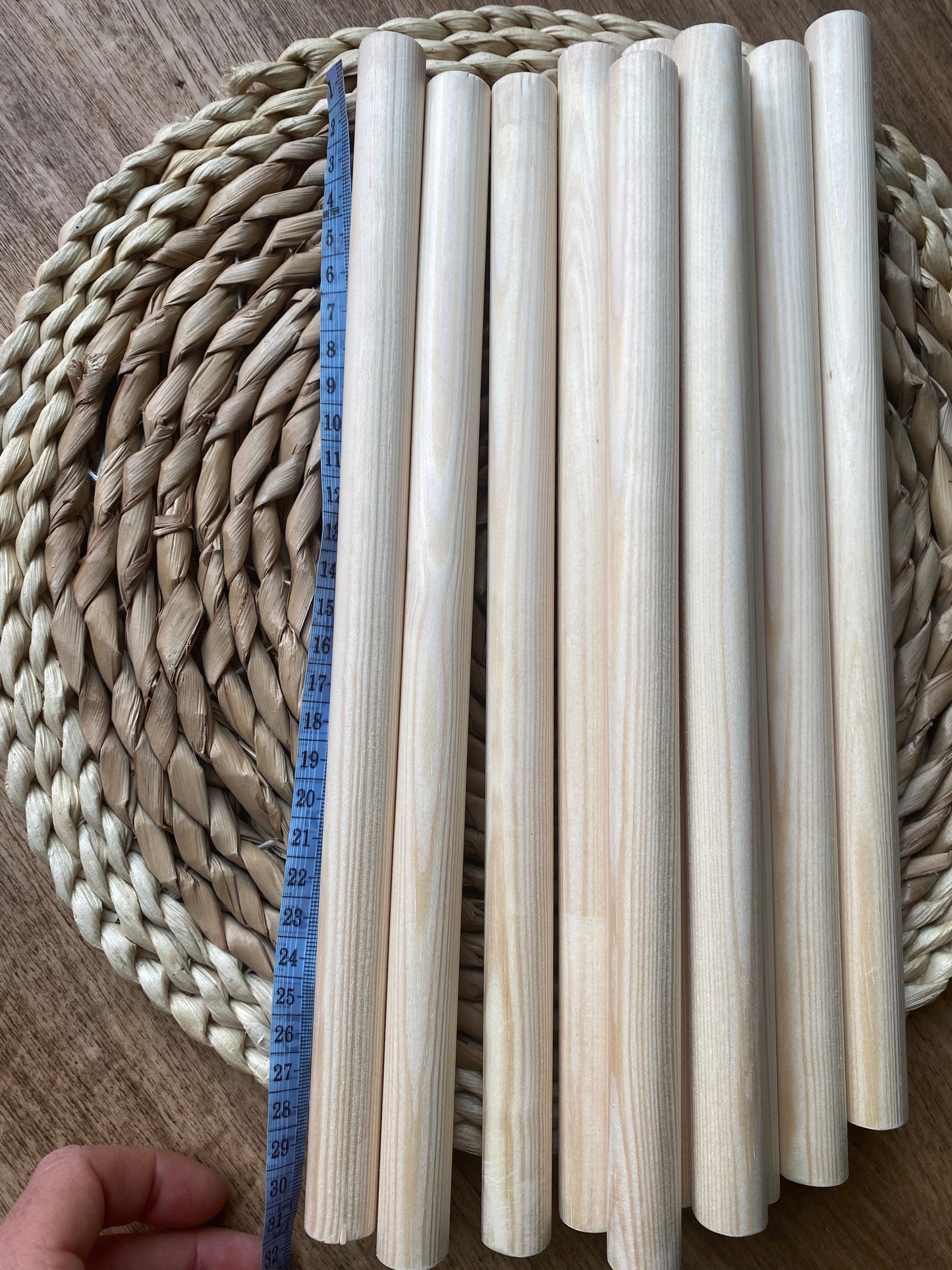 A pile of natural pine wooden dowels for macramé and crafts laid across a round wicker mat. A hand holds a thin purple tape measure next to the dowel, indicating a length of 30cm.