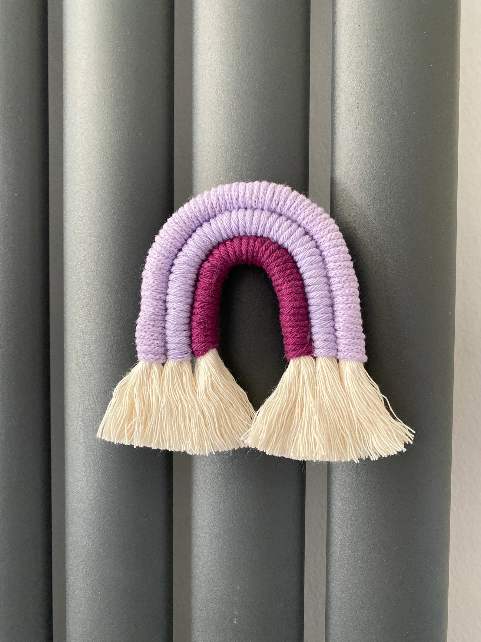 Small handmade macrame rainbow fridge magnet in soft cotton purple cords, on a dark grey radiator.