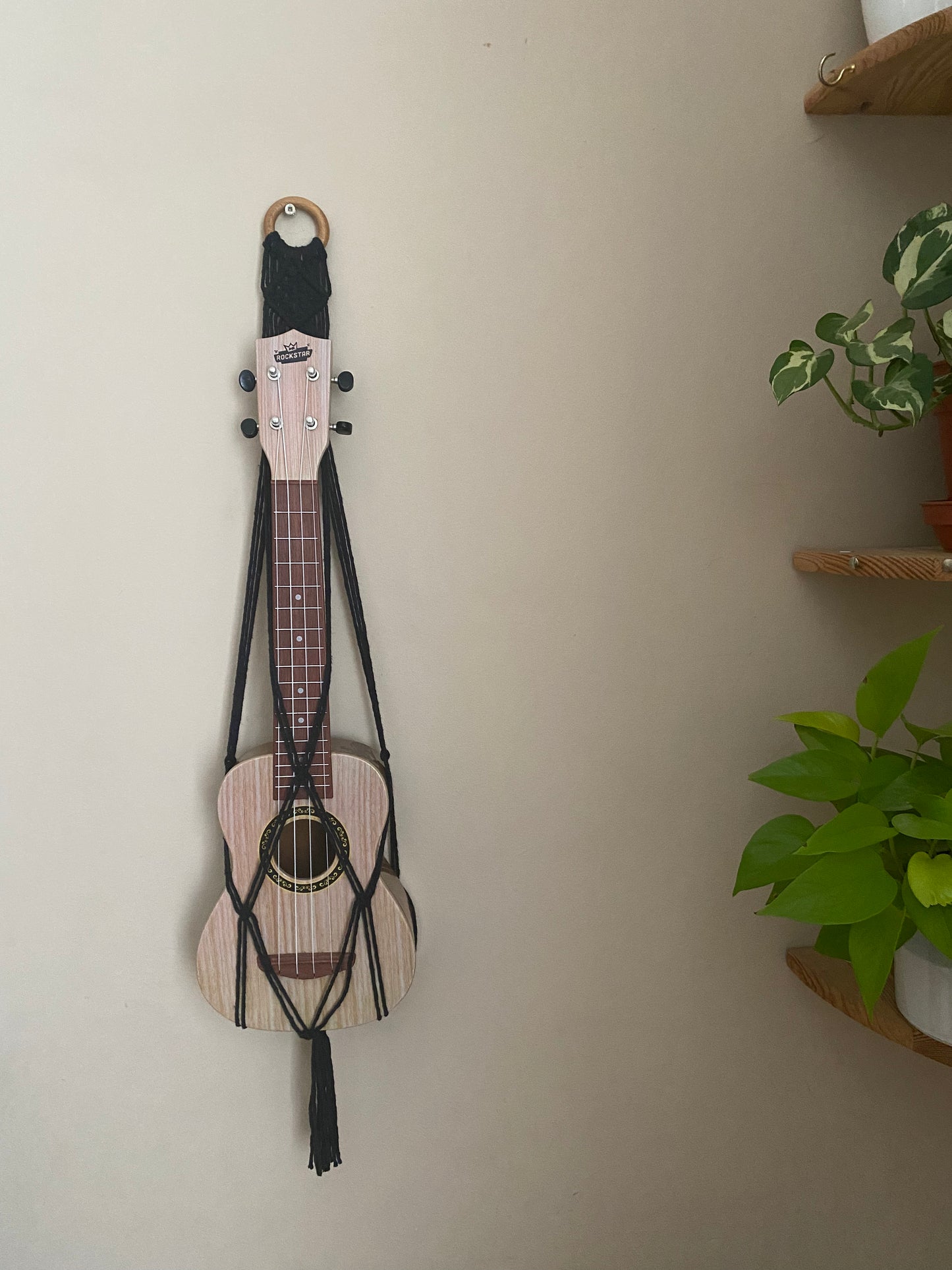 A light brown ukulele sits inside of a handmade macrame wall hanging ukulele hanger, which is made with black recycled cotton, and a repurposed wooden hanging ring. A green leafy plant is visible to the right of the image.
