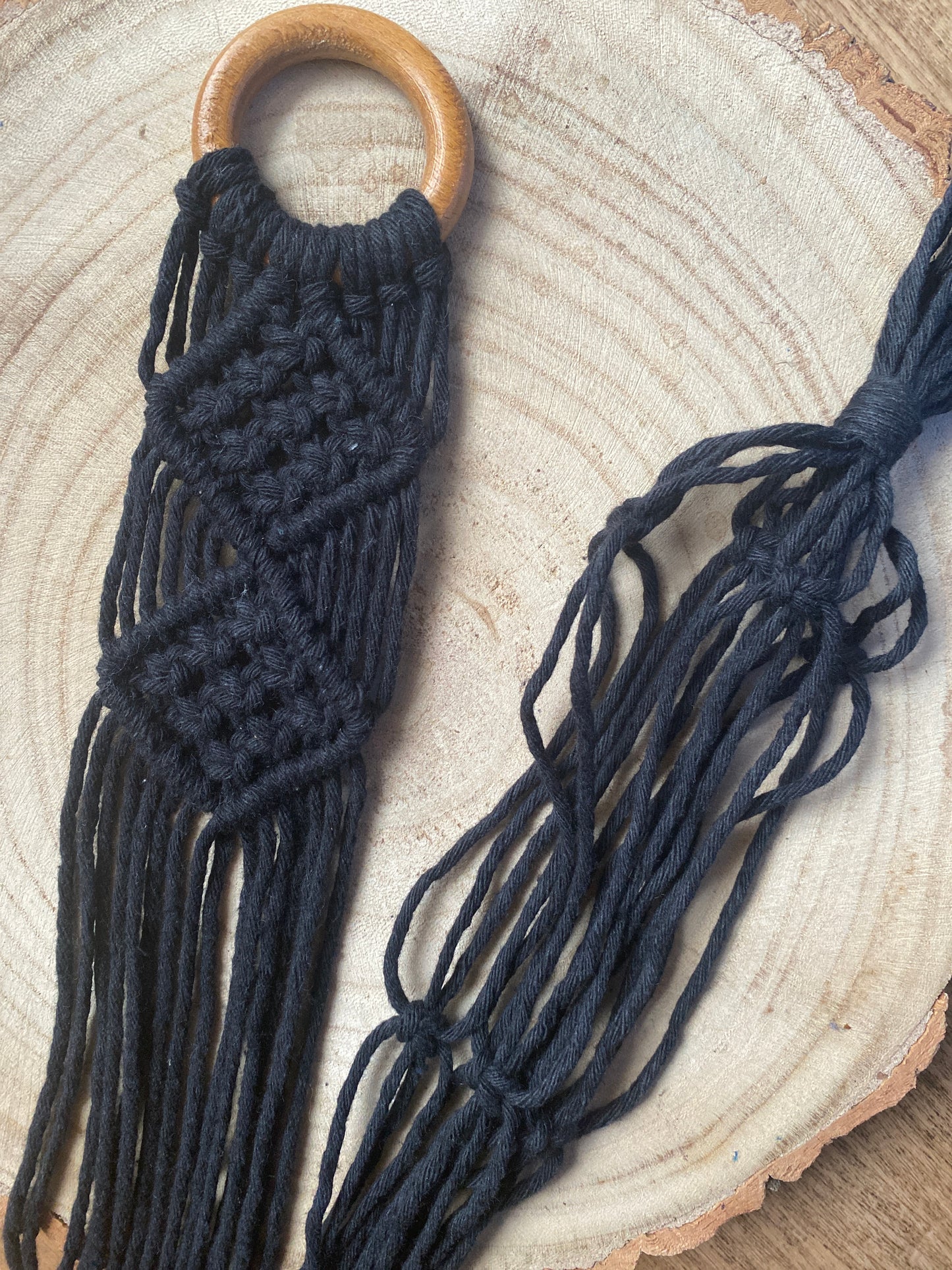 A handmade macrame ukulele wall hanger, made with black recycled cotton on a repurposed wooden hanging ring. The hanger is laid out on a round wooden surface, and you can see the intricate knot pattern.