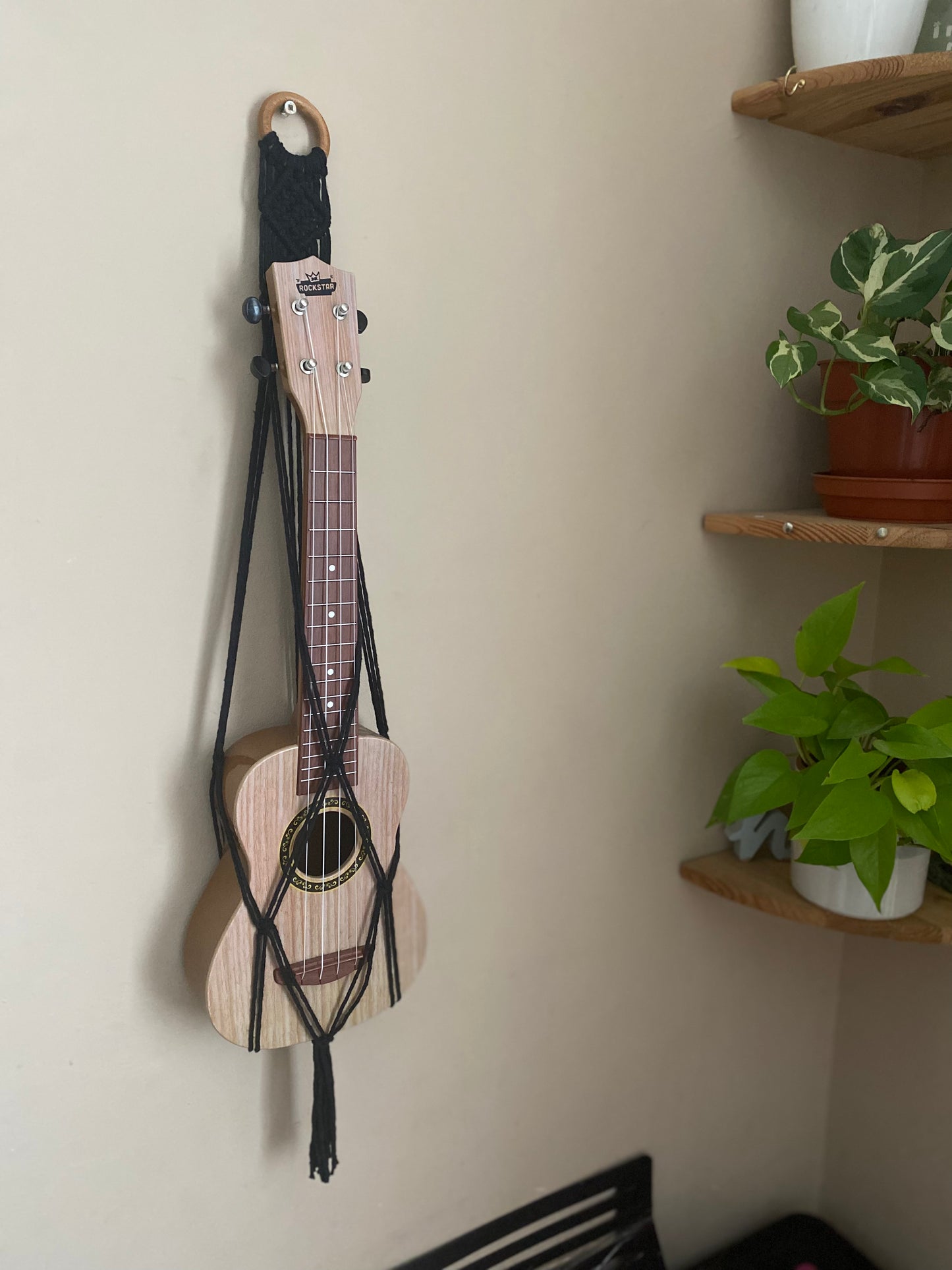 A light brown ukulele sits inside of a handmade macrame wall hanging ukulele hanger, which is made with black recycled cotton, and a repurposed wooden hanging ring. Two green plants sit on shelves next to the hanger.