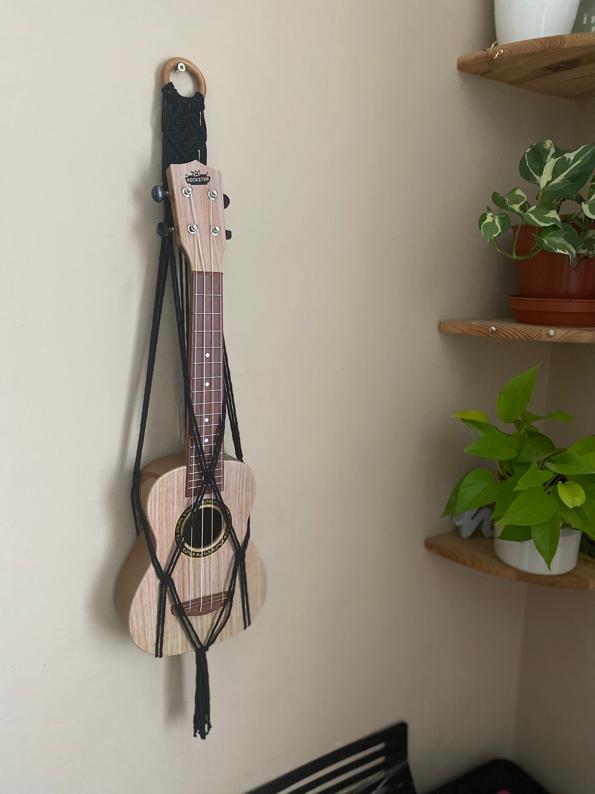 A light brown ukulele sits inside of a handmade macrame wall hanging ukulele hanger, which is made with black recycled cotton, and a repurposed wooden hanging ring. Two green plants sit on shelves next to the hanger.