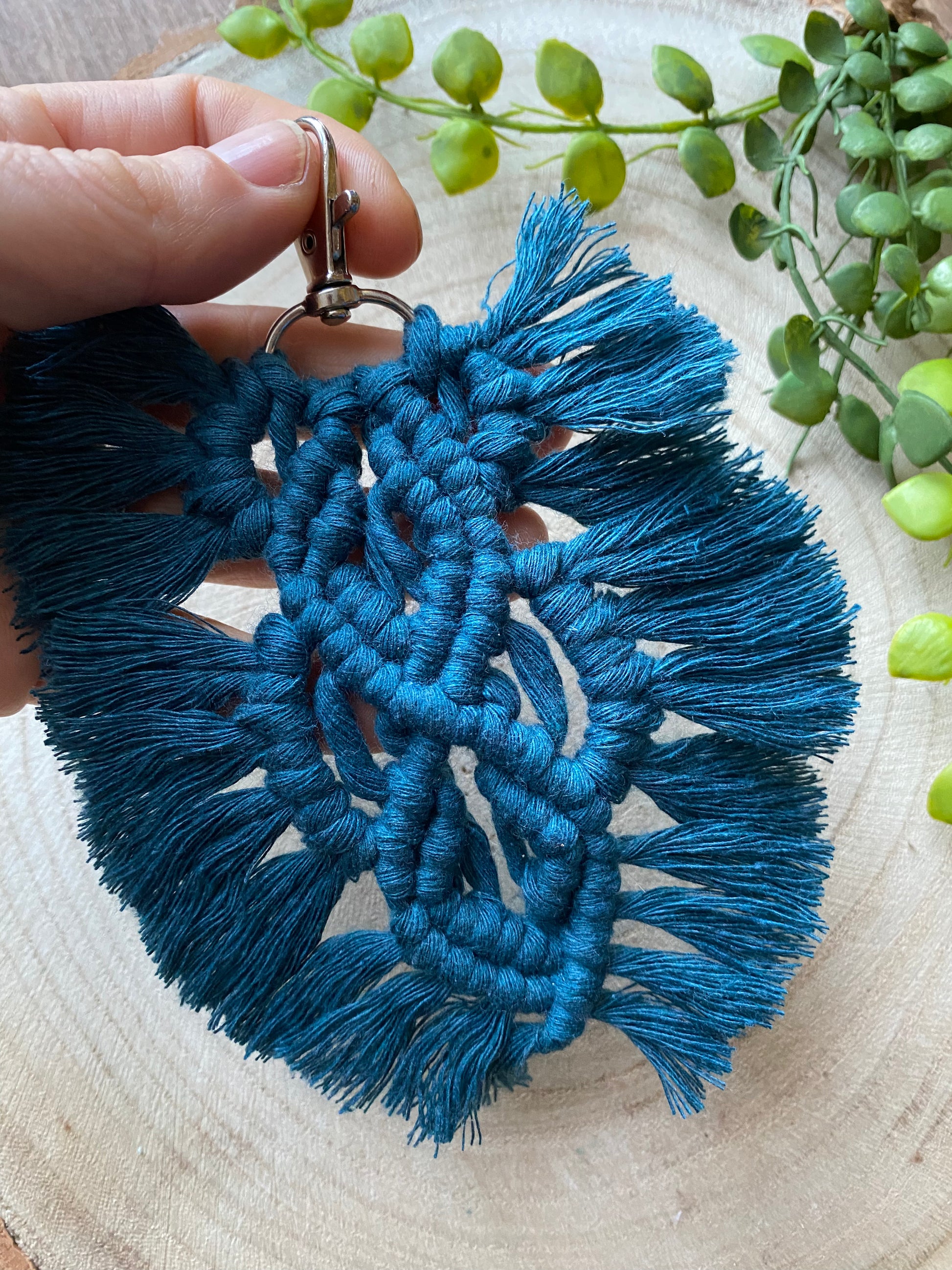 A hand holding a handmade macrame feather shaped keyring, made with peacock blue recycled cotton and a small metal clasp.