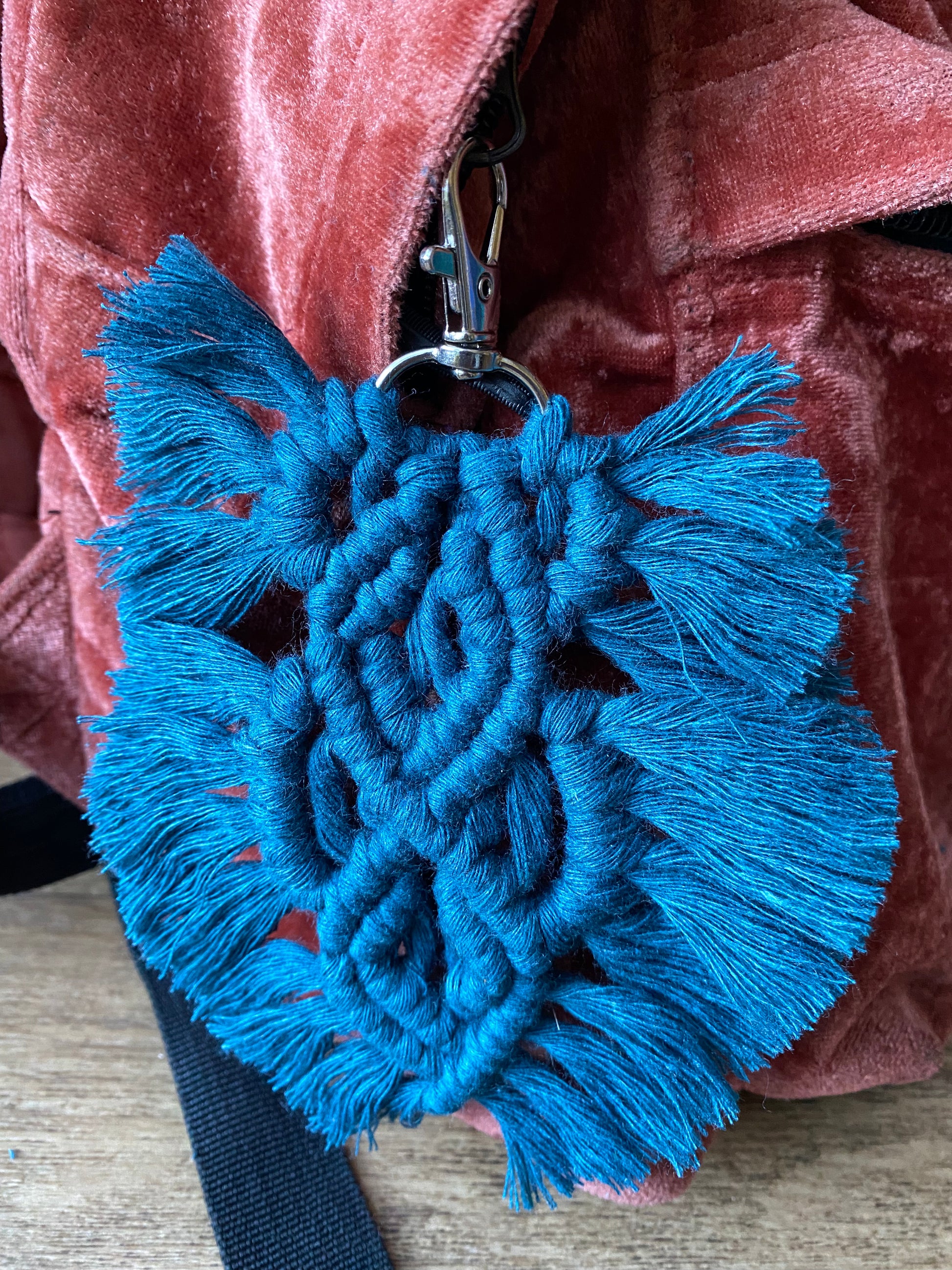 Handmade macrame feather shaped keyring, made with peacock blue recycled cotton and a small metal clasp, attached to the zip pull of a red velvet backpack.