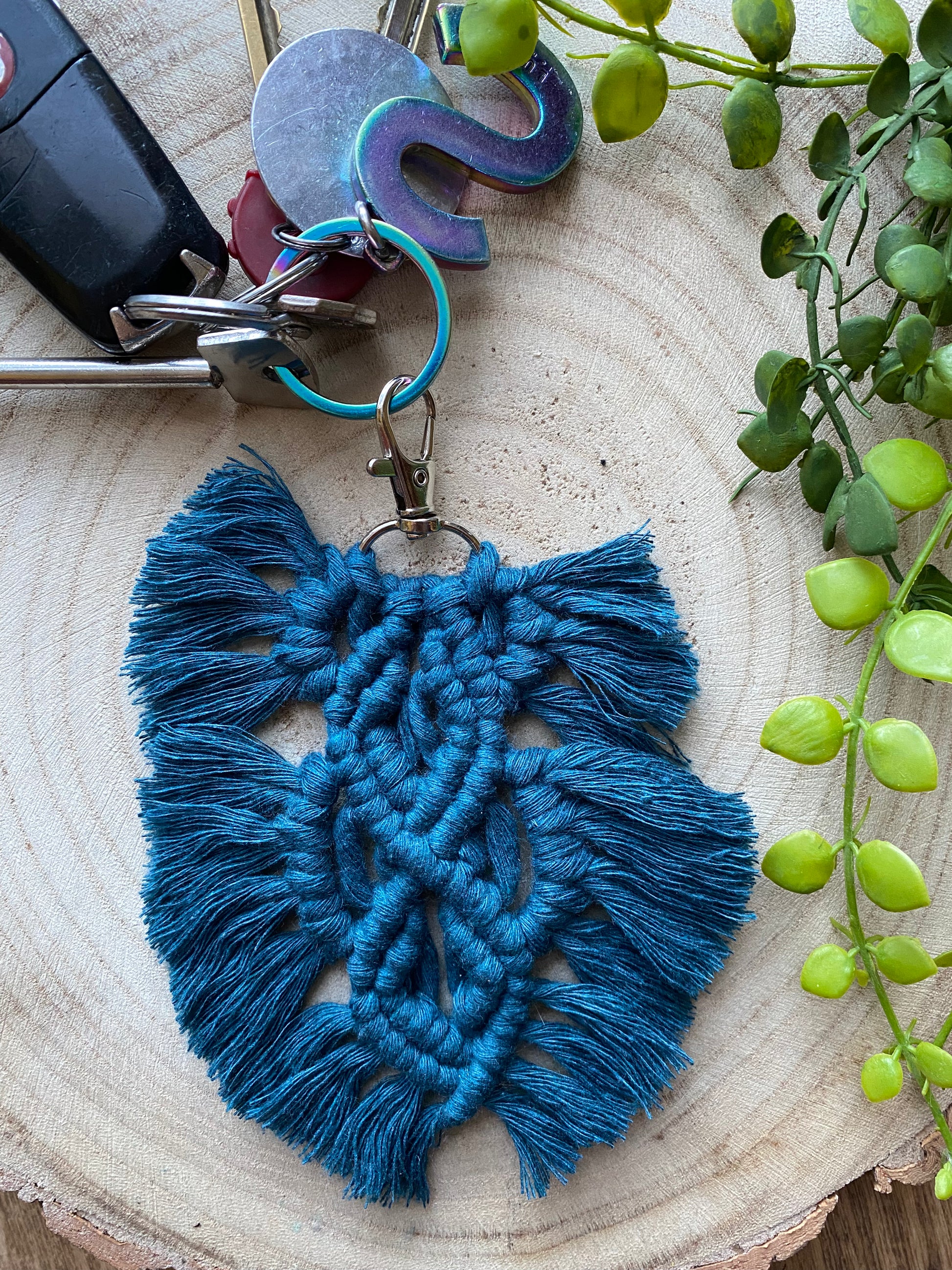 Handmade macrame feather shaped keyring, made with peacock blue recycled cotton and a small metal clasp, displayed on a wooden surface, attached to a small set of keys.