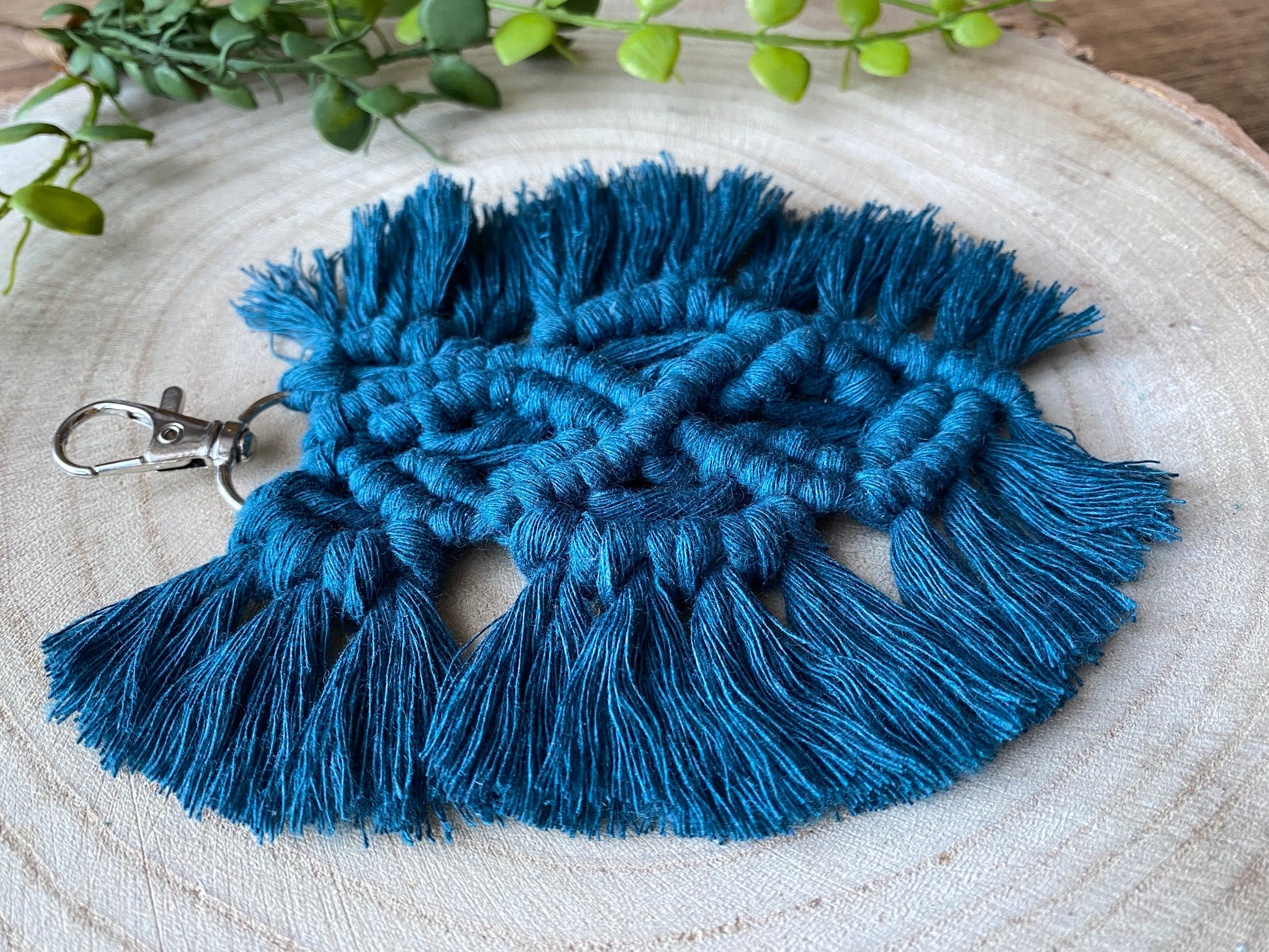 Close up of a handmade macrame feather shaped keyring, made with peacock blue recycled cotton with a small metal clasp, on a round wooden surface.