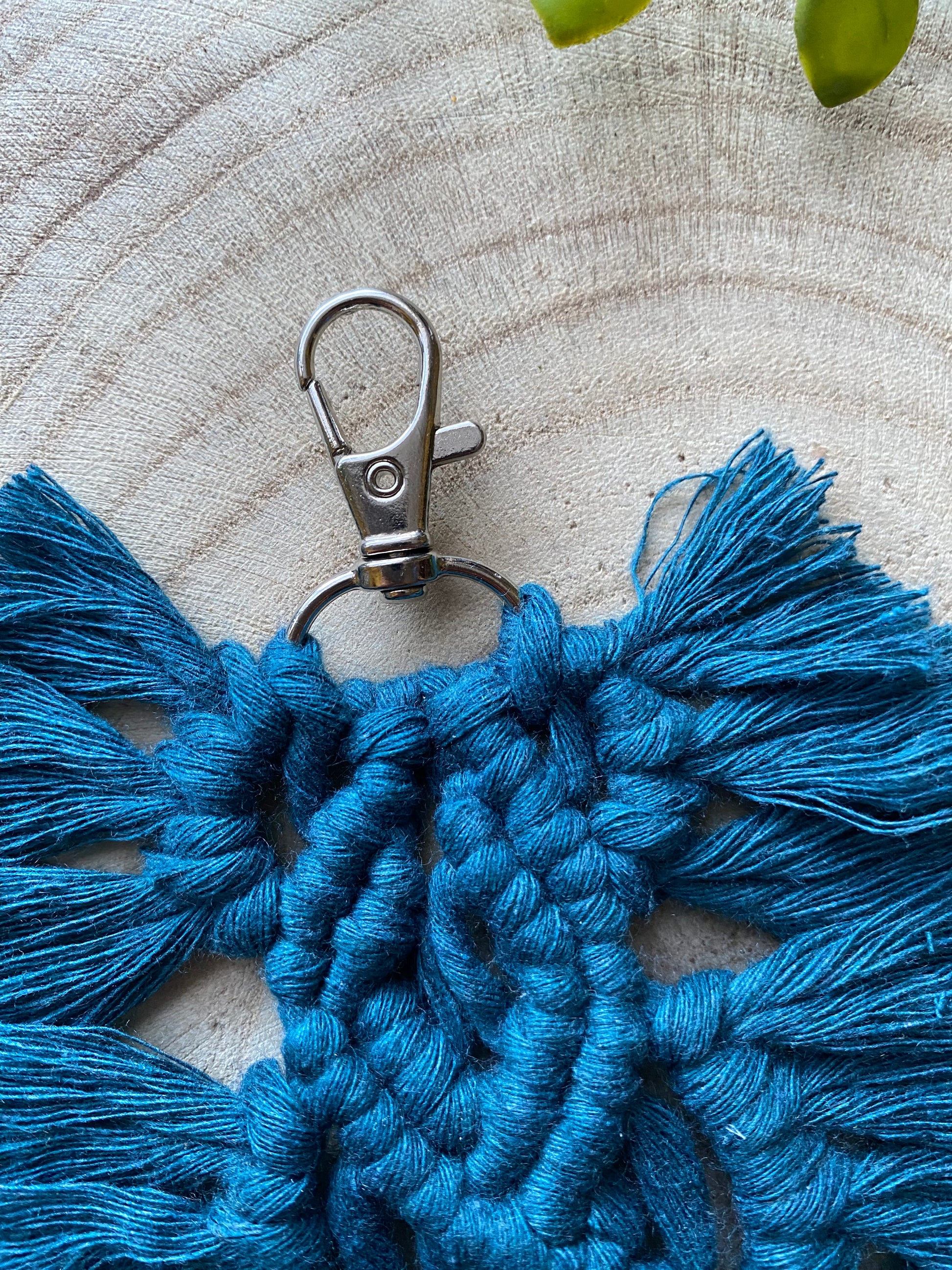 Close up view of the top part of a handmade macrame feather shaped keyring, made with peacock blue recycled cotton and a small metal clasp.