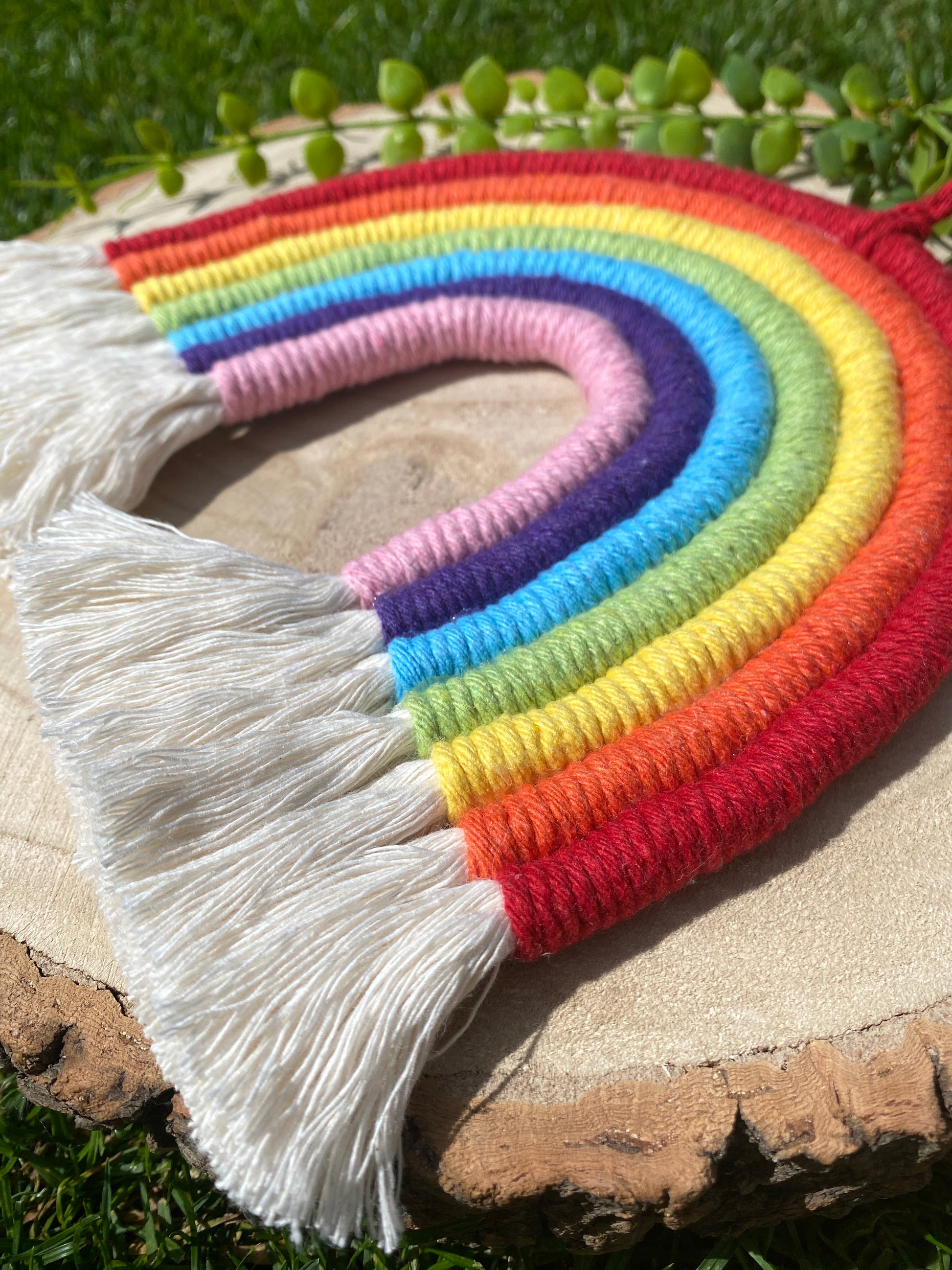 A handmade macrame rainbow, made with 7 arches of colourful yarn, with white tassels at the bottom and a small hanging loop at the top. Displayed on a round wooden board.