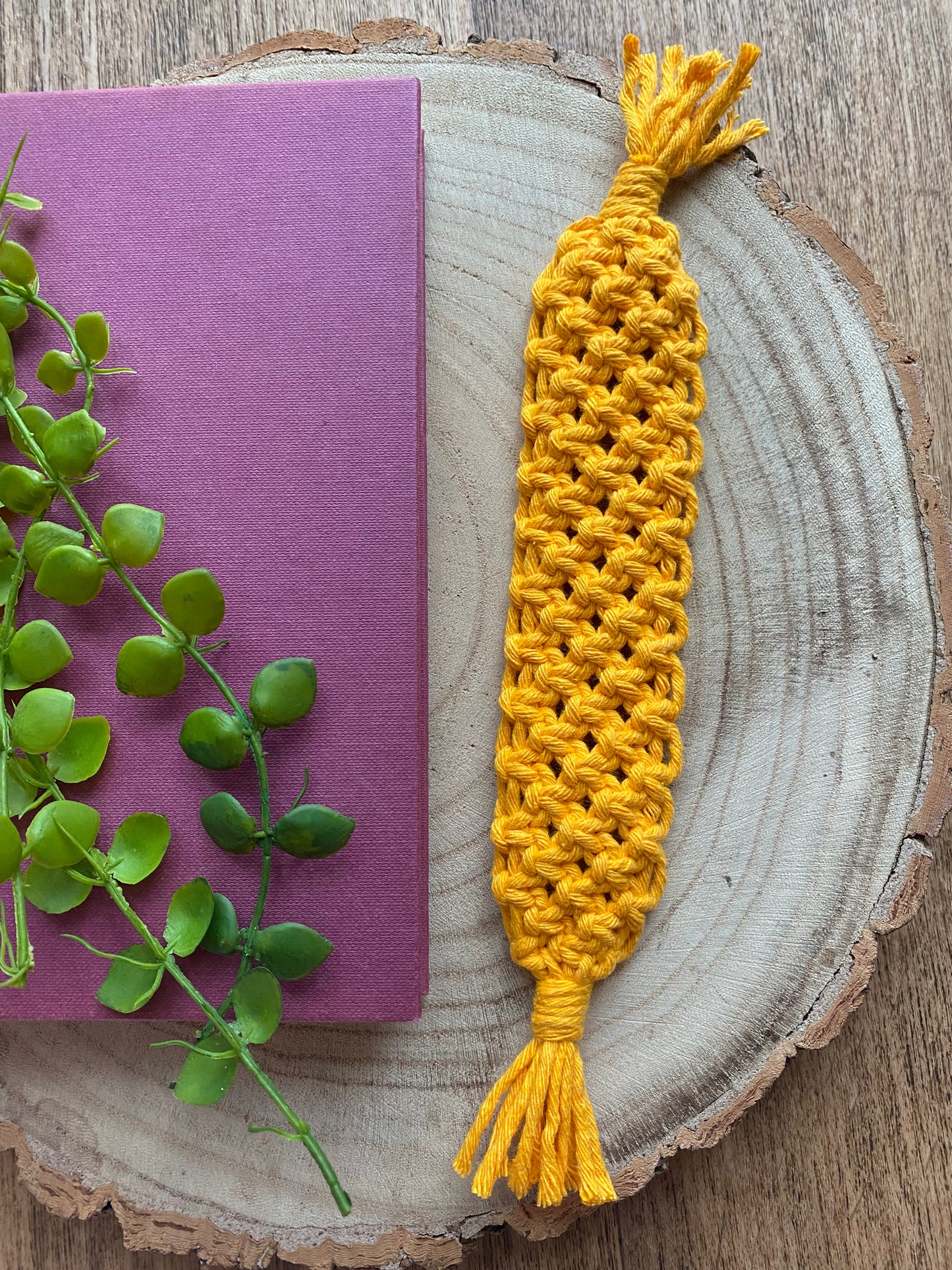 Handmade macramé bookmark with knotted design made with bright amber yellow recycled cotton, laid beside a hardback book on a wooden surface.