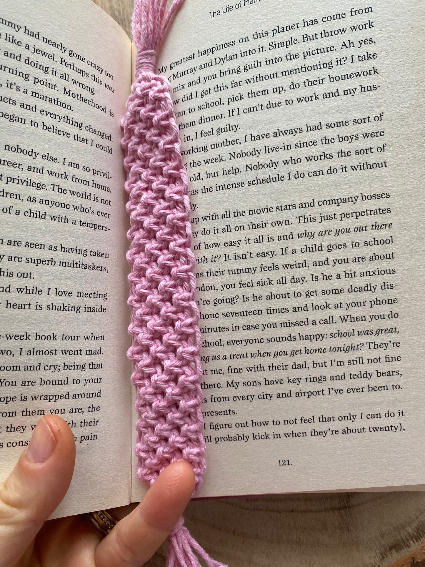 Handmade macramé bookmark with knotted design made with baby pink recycled cotton, laid across an open hardback book, which is being held in a person's hand at the bottom.