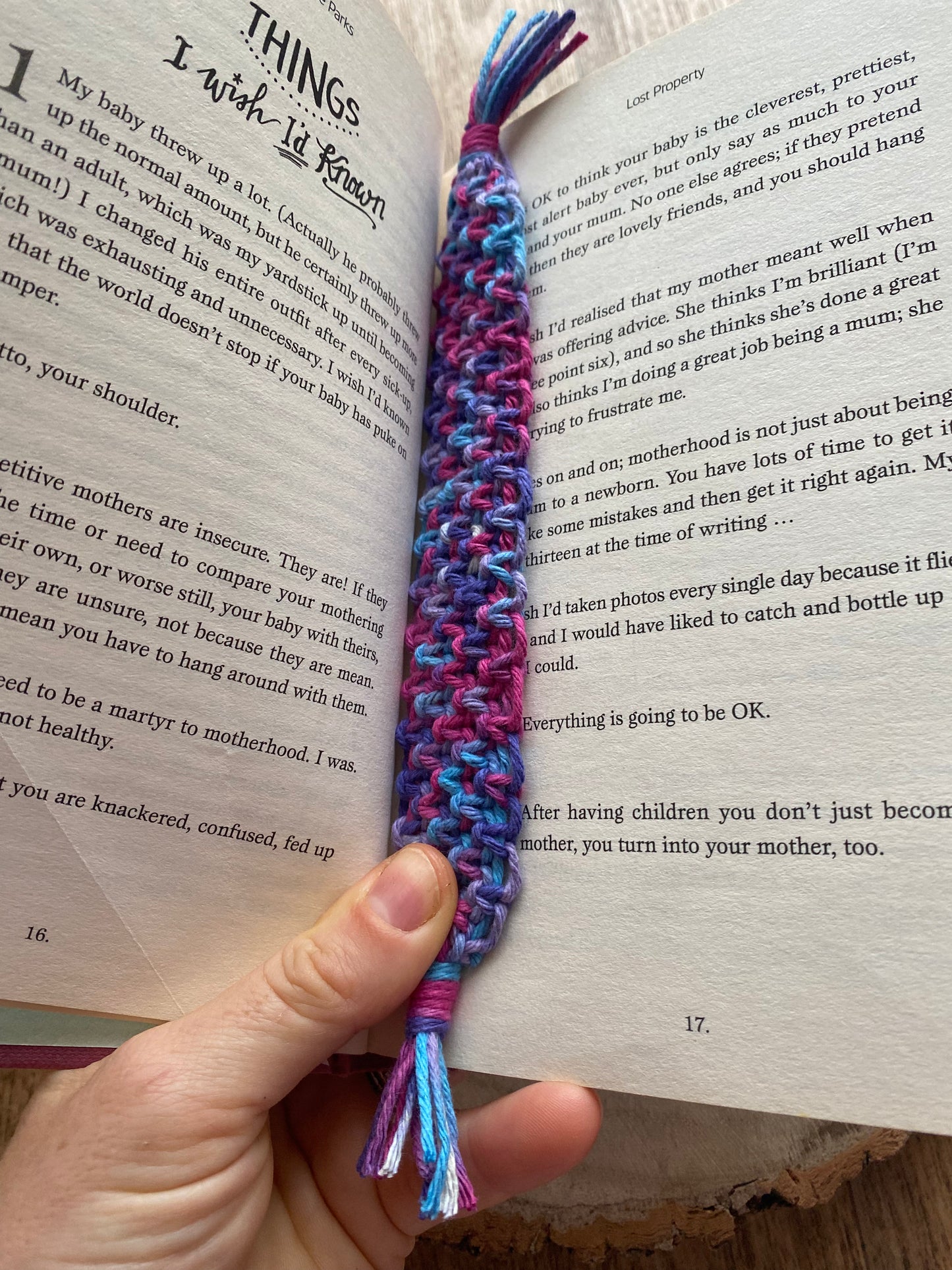 Handmade macramé bookmark with knotted design made with purple recycled cotton, laid inside an open hardback book which is held by a person's hand at the base.