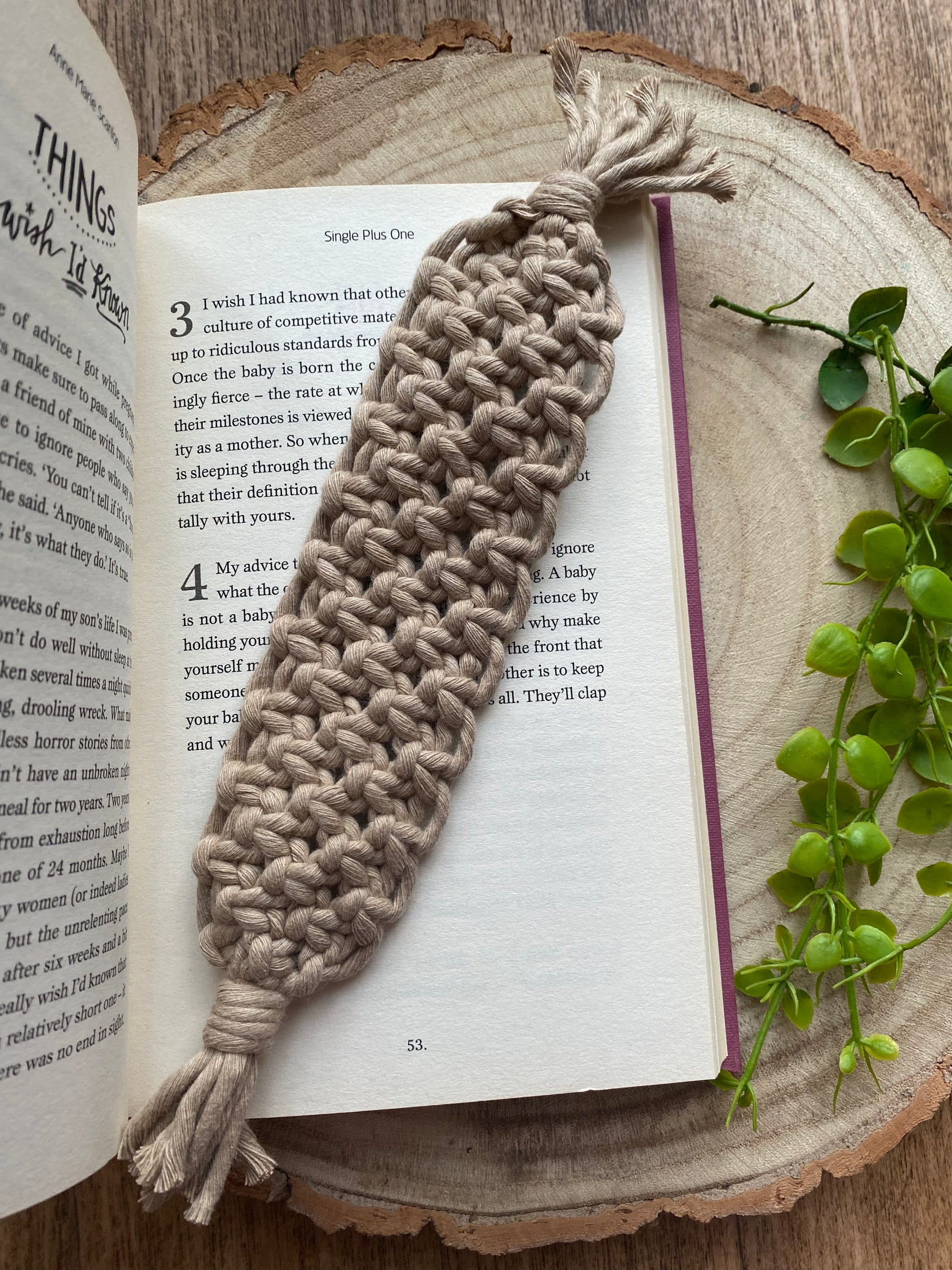 Handmade macramé bookmark with knotted design made with beige recycled cotton, laid across an open hardback book on a wooden surface.