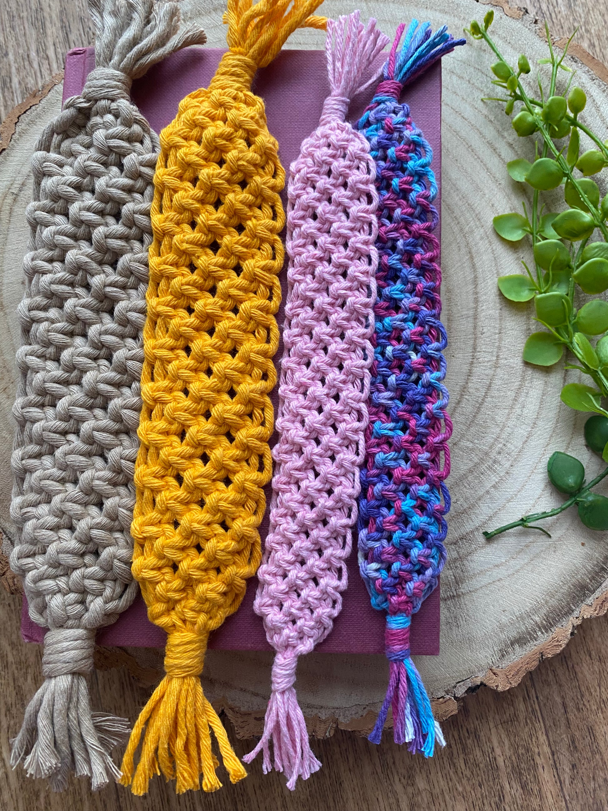 Four handmade macramé bookmarks with knotted design in a variety of bright colours, laid across a red hardback book on a wooden surface.