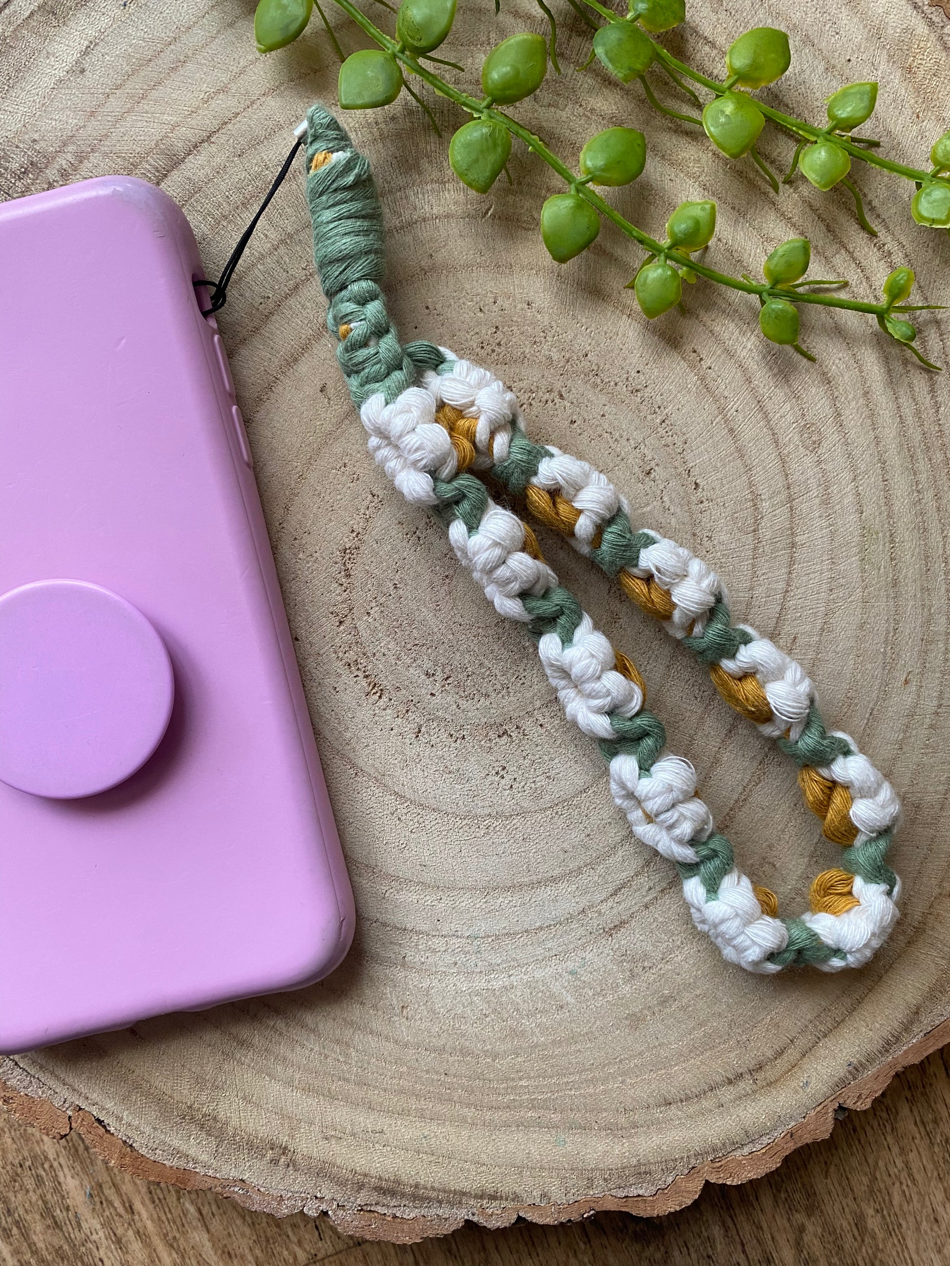 Handmade macrame daisy chain phone charm wristlet, made with white, yellow and green yarns, attached to a pink phone case with pop socket, and laid on a wooden surface.
