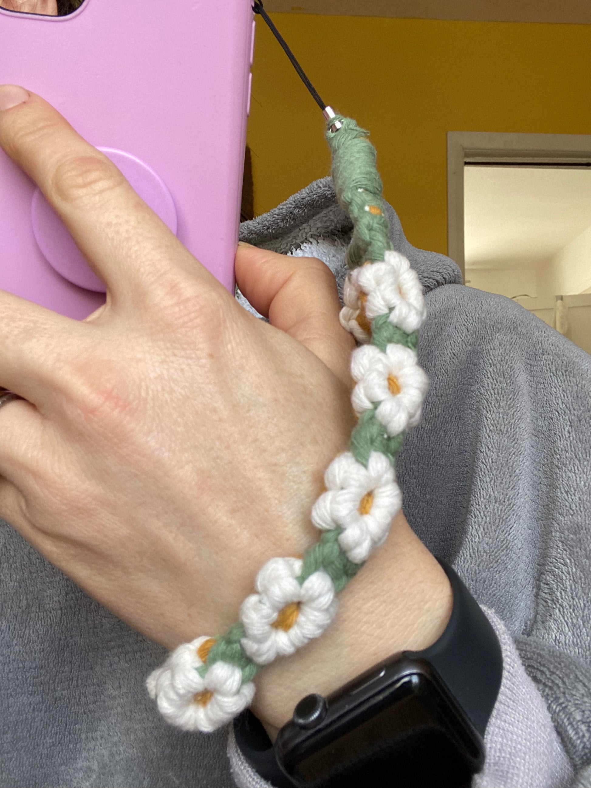 A woman holding a pink phone case, with a daisy chain phone charm wristlet attached to it, around her wrist. The woman wears a fluffy grey hoody and has a black watch on her wrist.