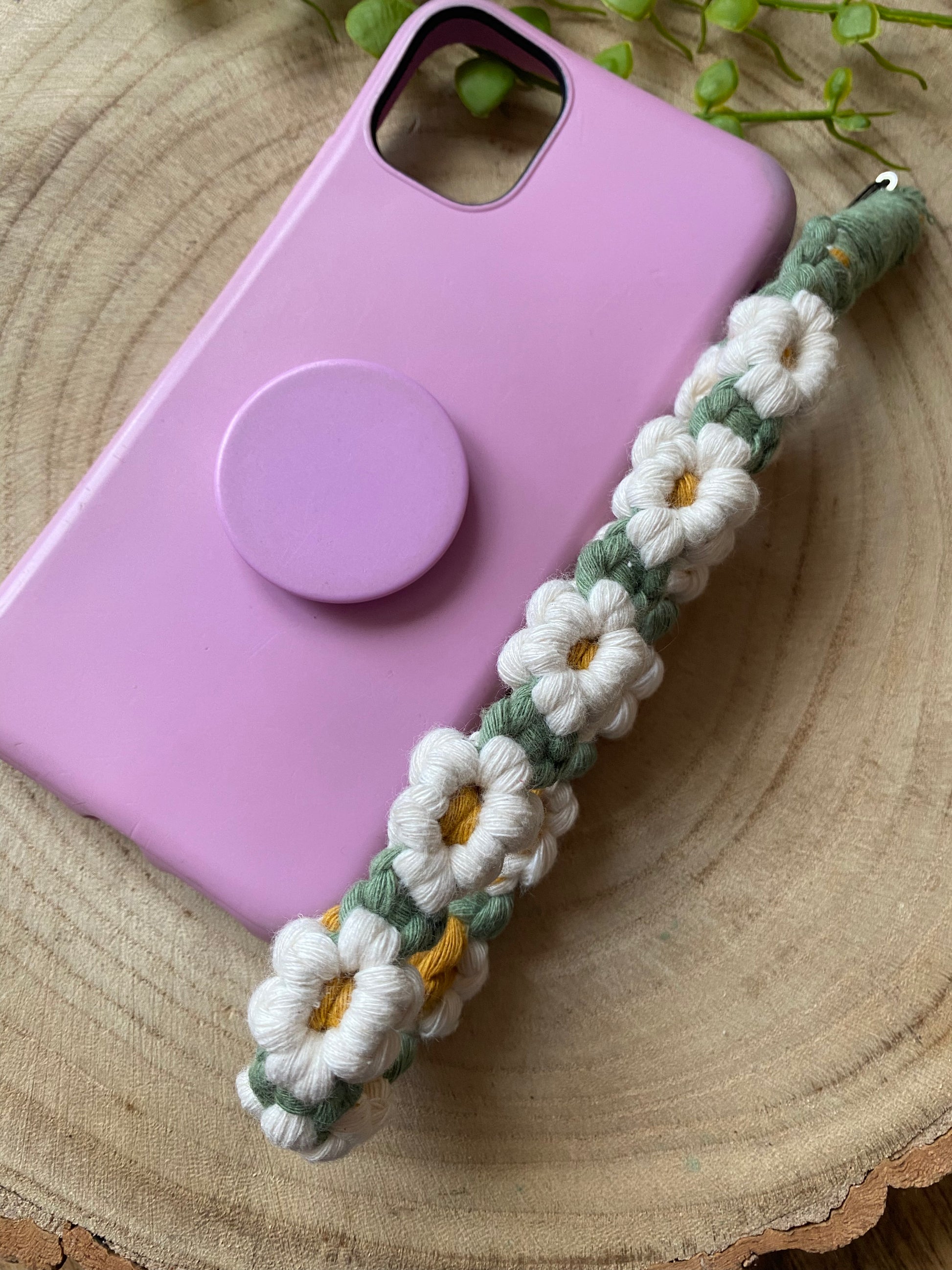 Handmade macrame daisy chain phone charm wristlet, made with white, yellow and green yarns, attached to a pink phone case with pop socket, and laid on a wooden board.
