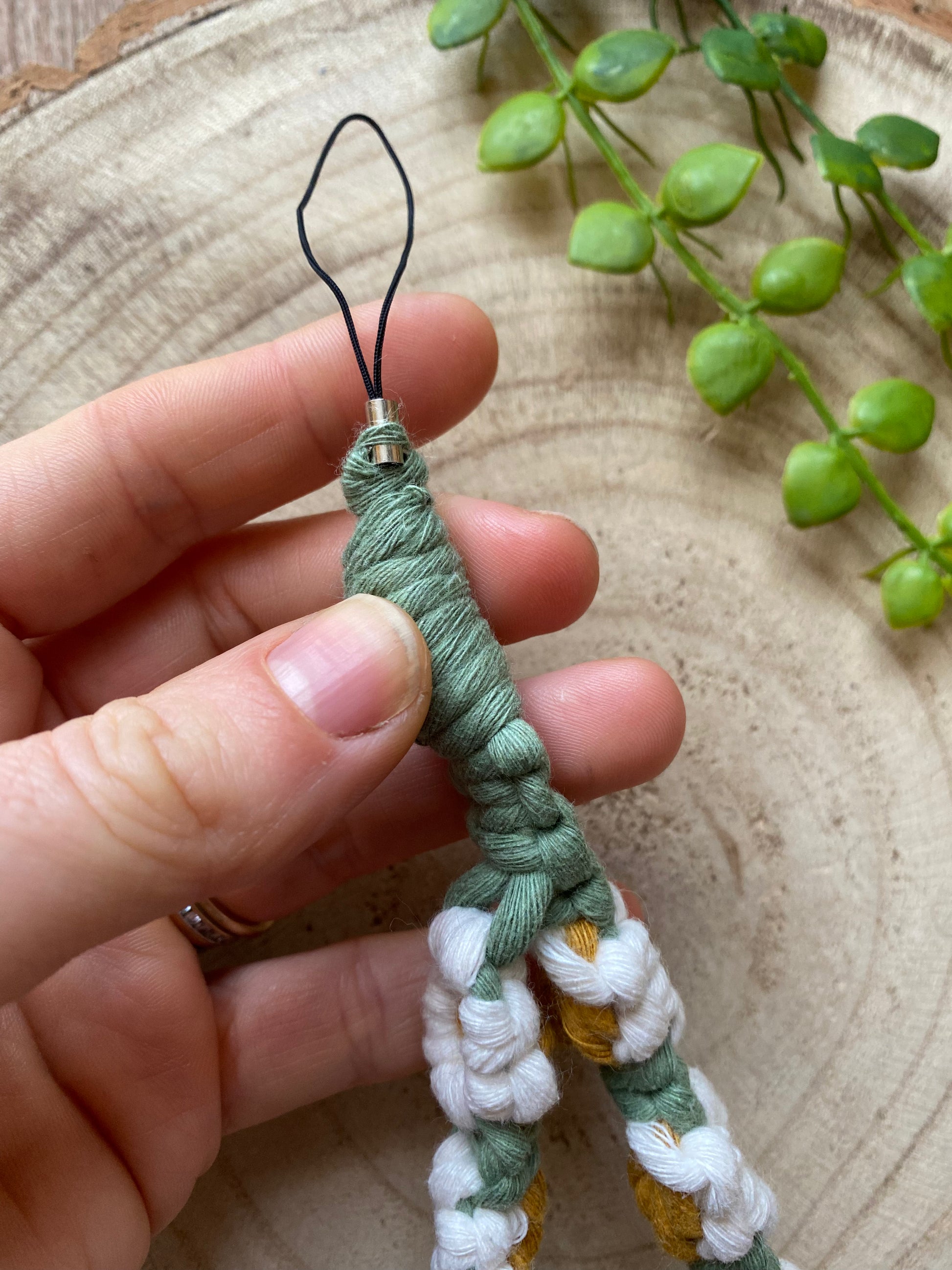 A hand holding the end of a macrame daisy chain phone charm wristlet, made with white, yellow and green yarns.