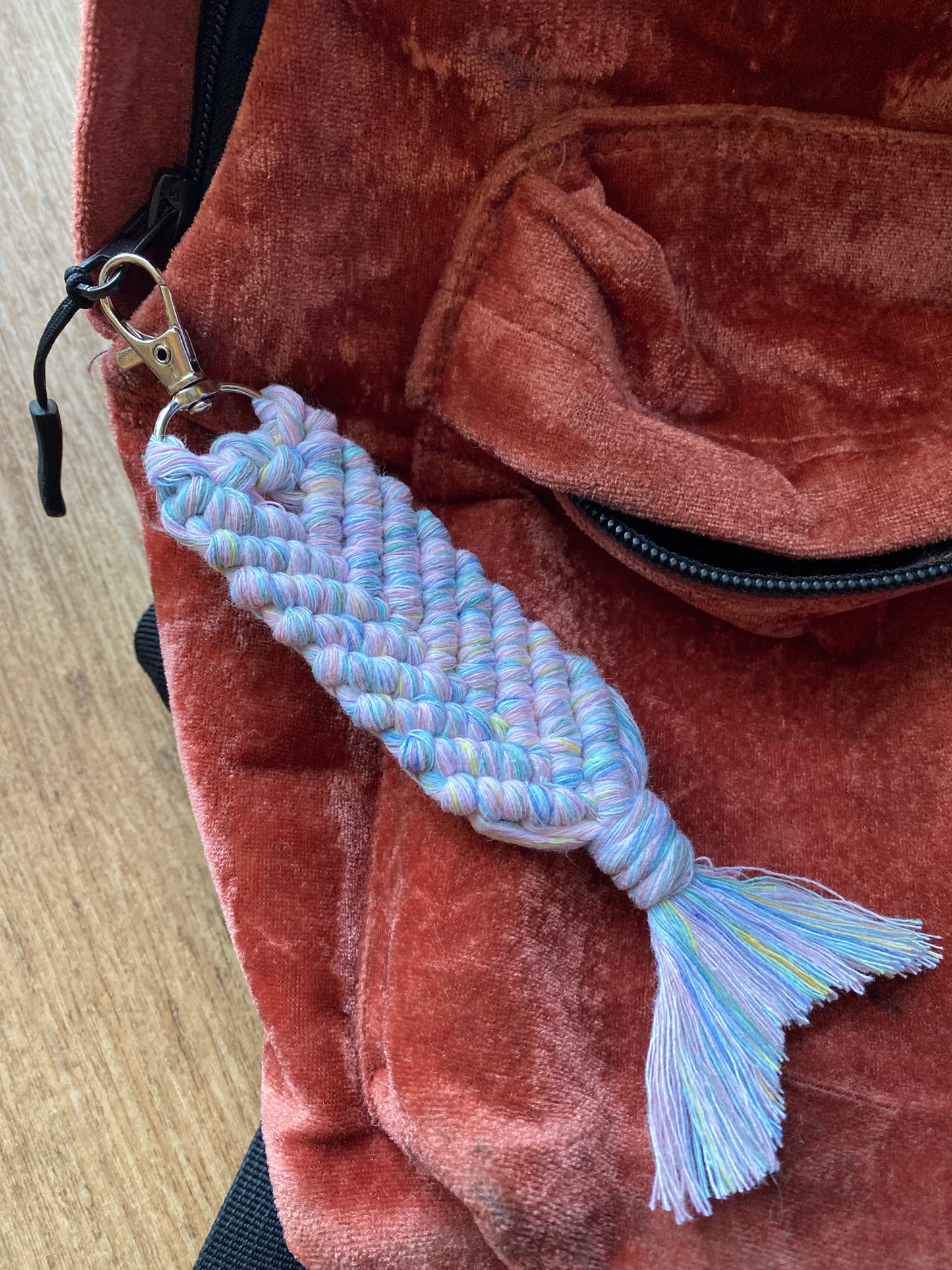 A handmade macrame keyring, knotted using soft pastel coloured recycled cotton in the shape of a mermaid's tail, and finished with a metal lobster clasp. Shown attached on to the zipper of a velvet red backpack.