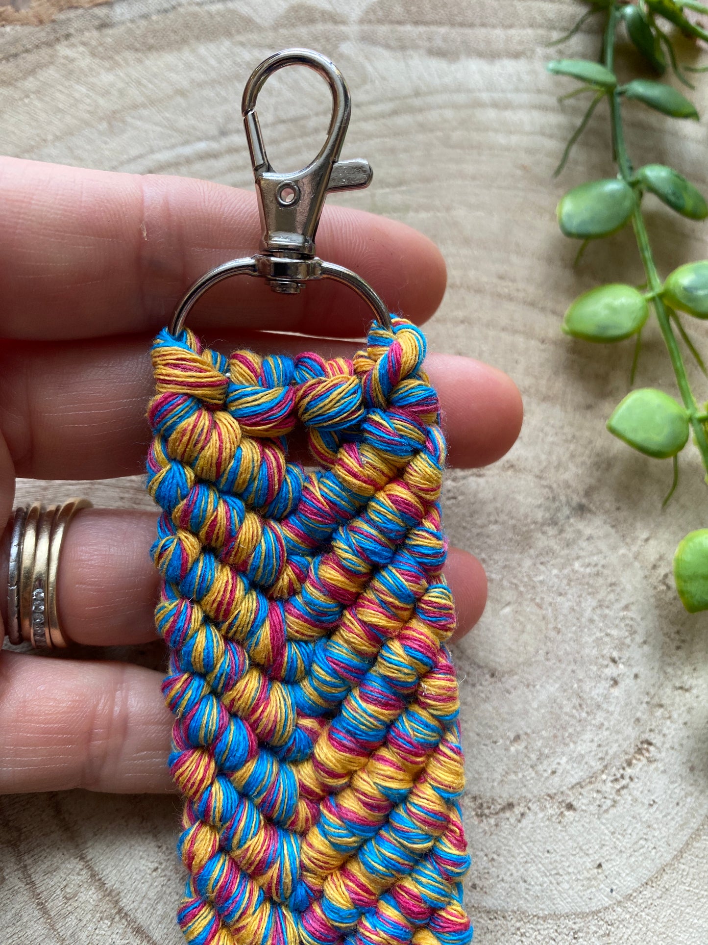 A hand holding a handmade macrame keyring, knotted using brightly coloured recycled cotton and finished with a metal lobster clasp.