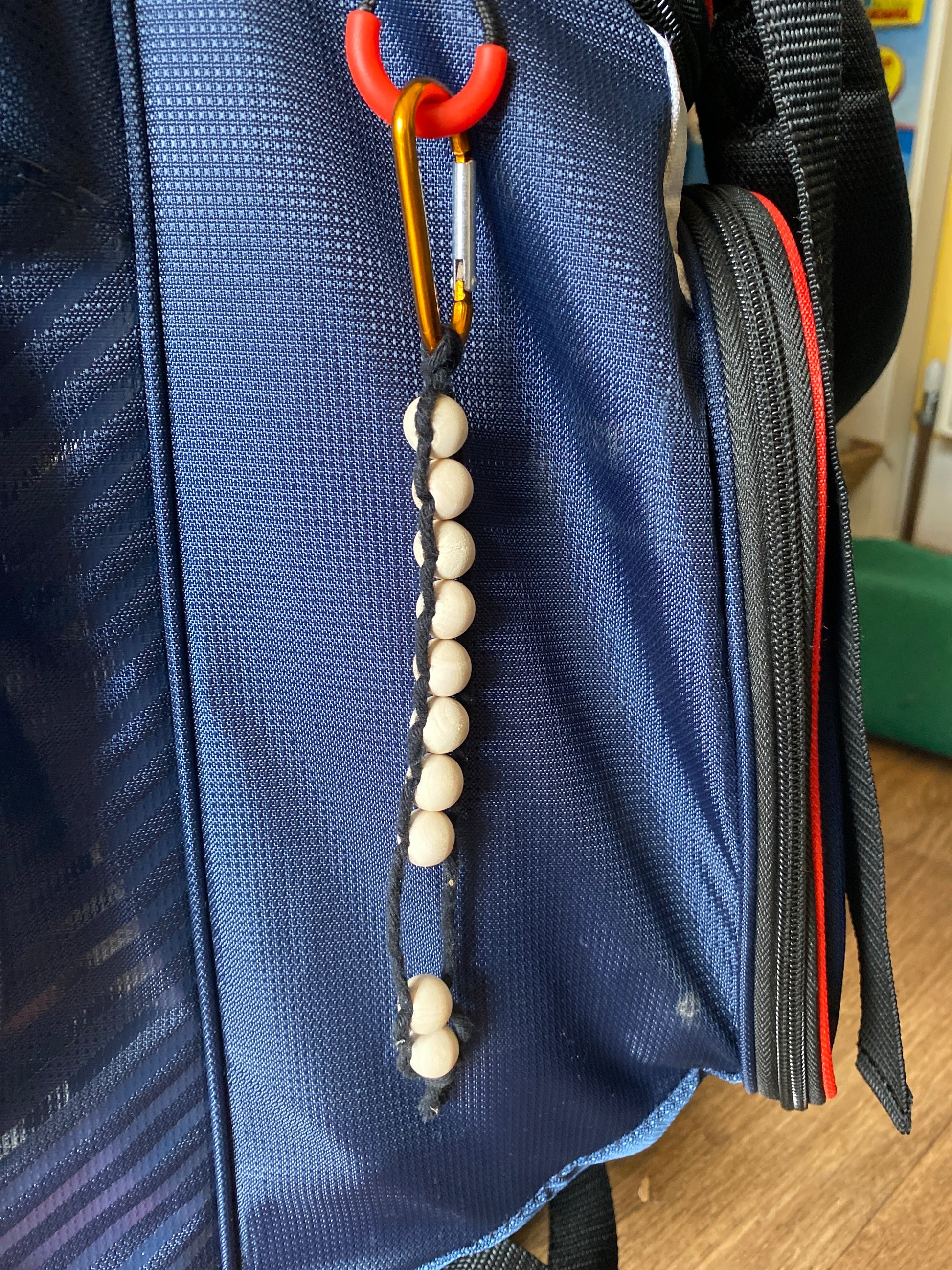 A handmade macramé golf stroke counter made with black yarn and 10 small plain wooden beads, attached to a blue golf bag with an orange carabiner clip.