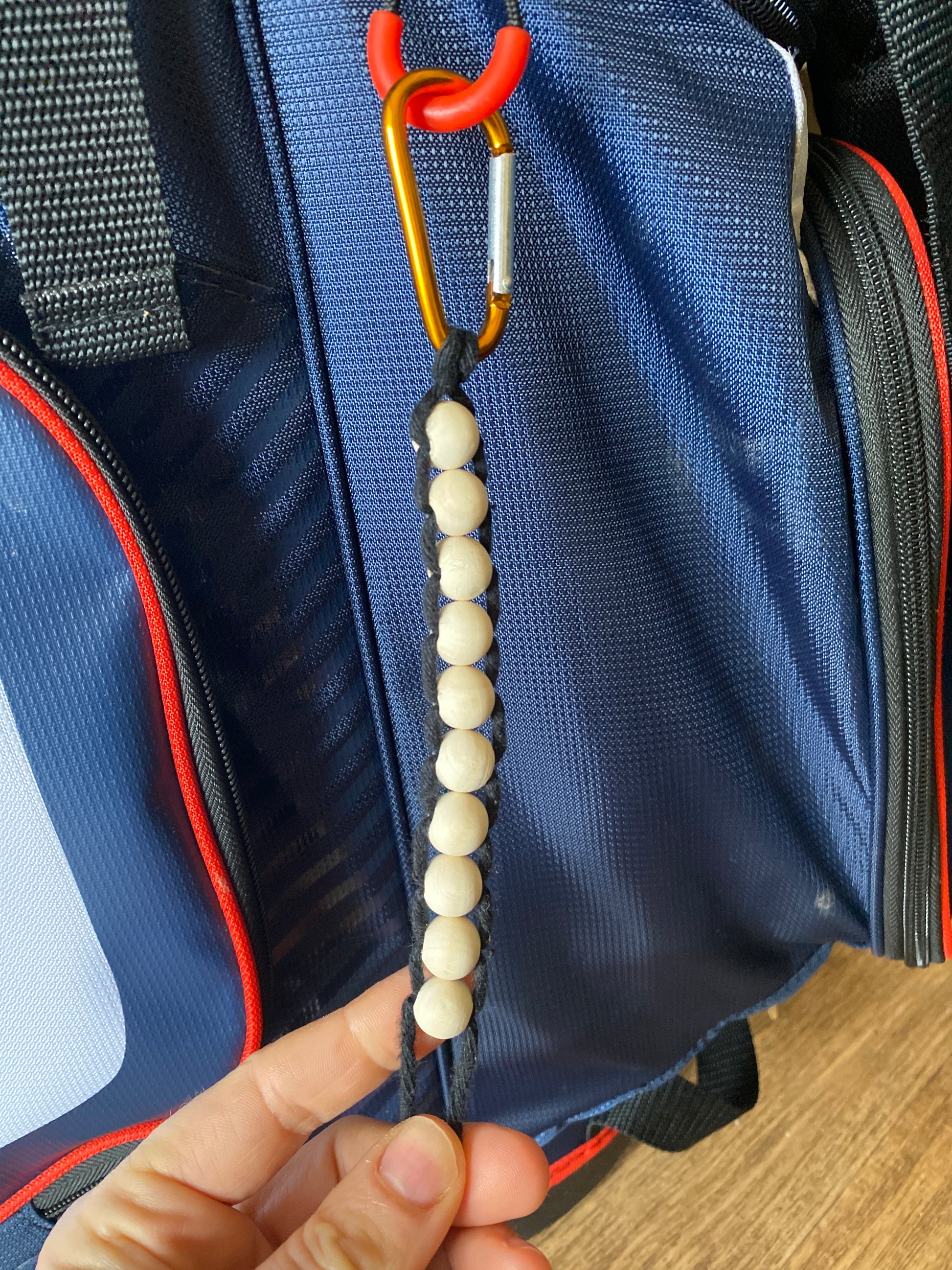 A hand holds a handmade macramé golf stroke counter made with black yarn and 10 small plain wooden beads, attached to a blue golf bag with an orange carabiner clip.