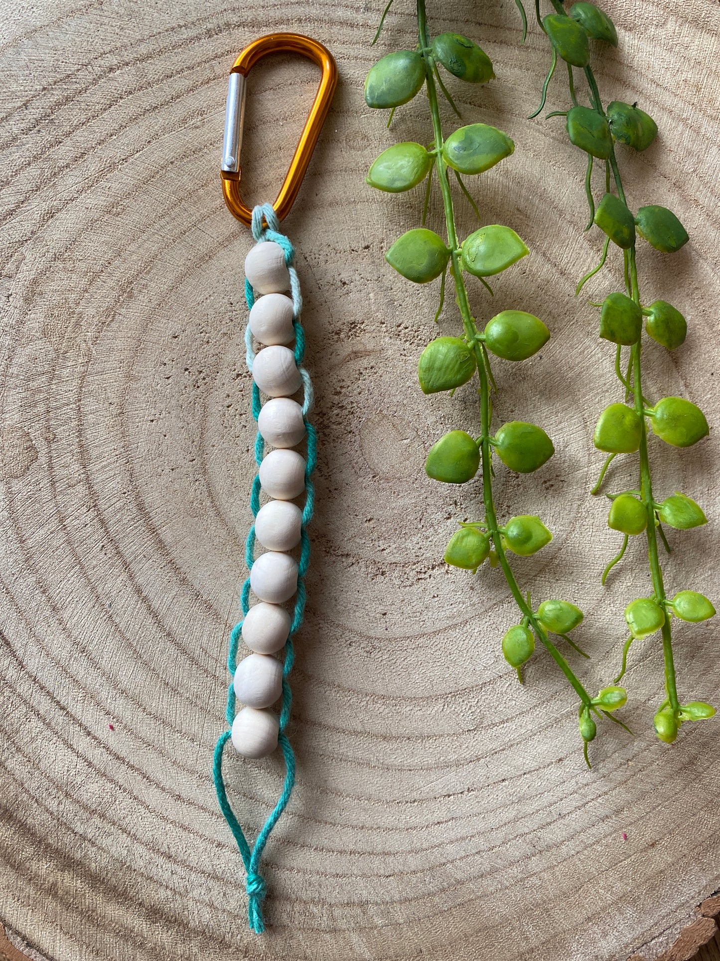 A handmade macramé golf stroke counter made with mint green yarn and 10 small plain wooden beads, laid out on a wooden surface with some plant vines.