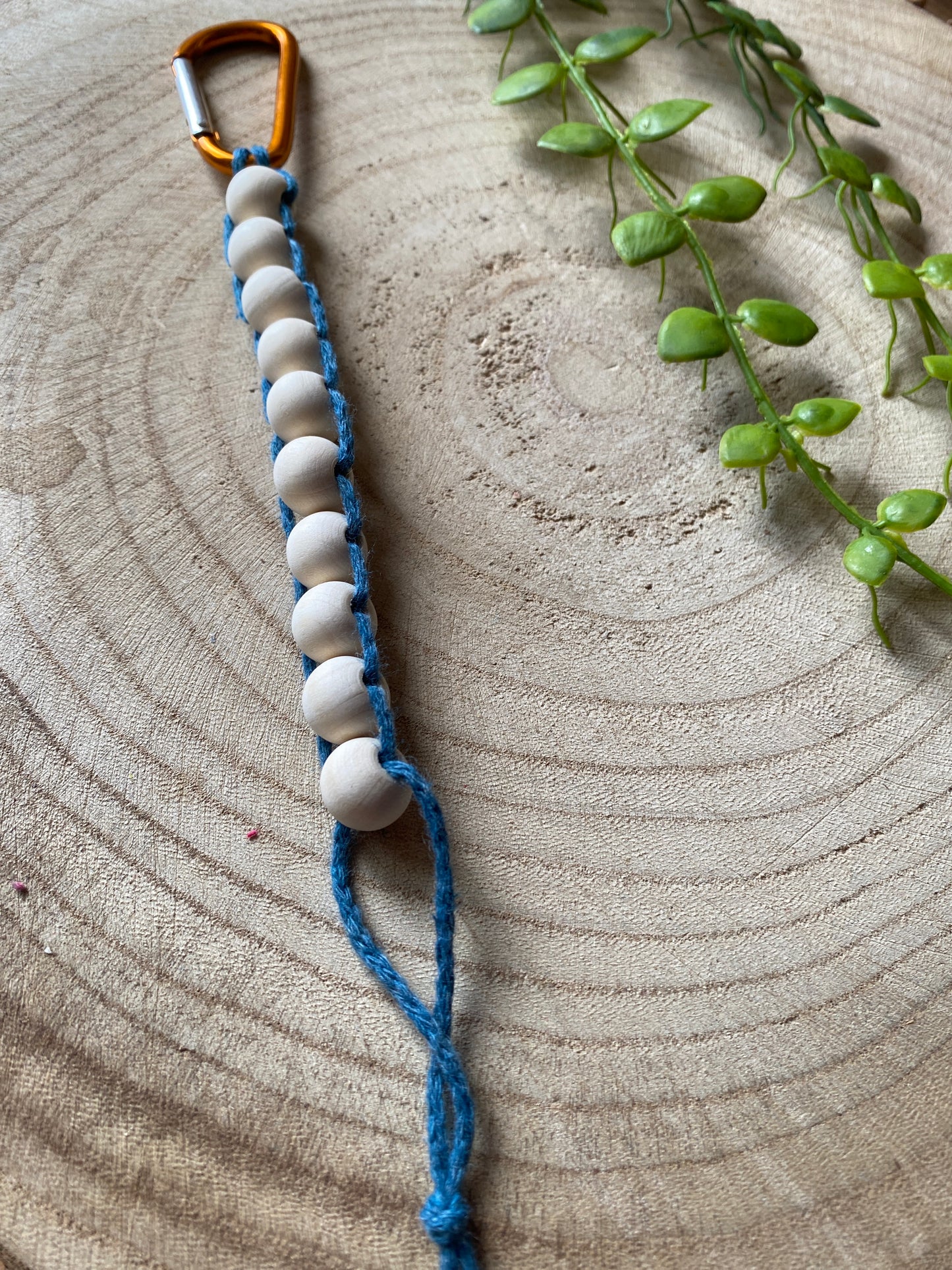 A close up image of a handmade macramé golf stroke counter made with teal blue yarn and 10 small plain wooden beads, attached to an orange carabiner clip.