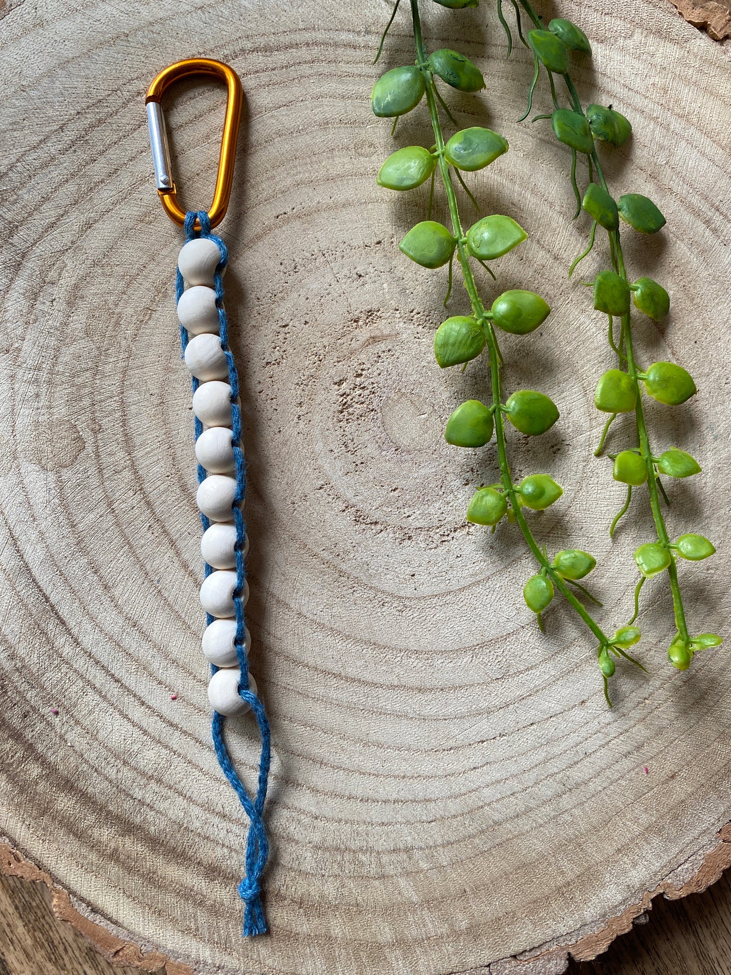 A handmade macramé golf stroke counter made with teal blue yarn and 10 small plain wooden beads, on an orange carabiner clip.
