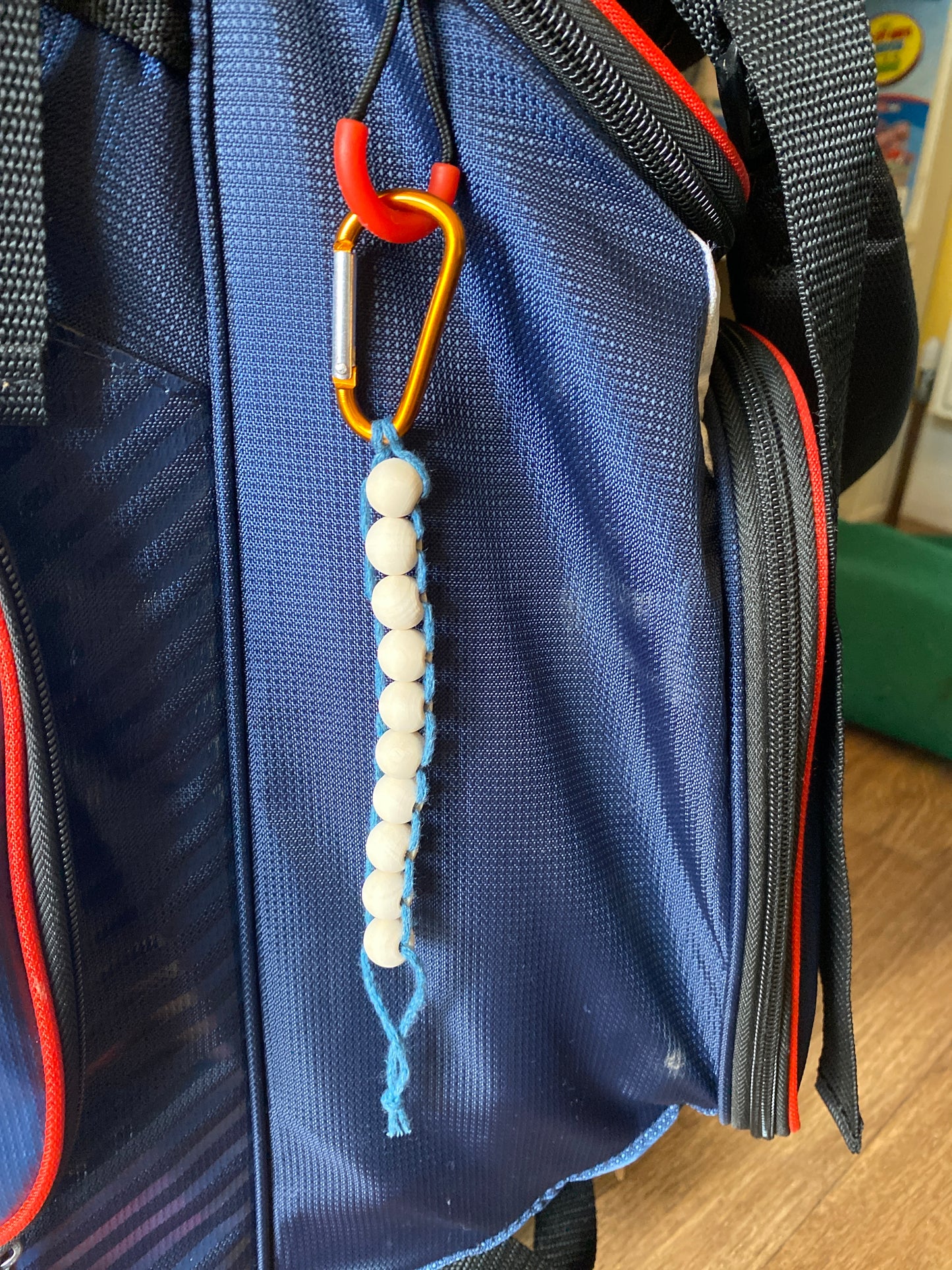 A handmade macramé golf stroke counter made with teal yarn and 10 small plain wooden beads, attached to a blue golf bag with an orange carabiner clip.