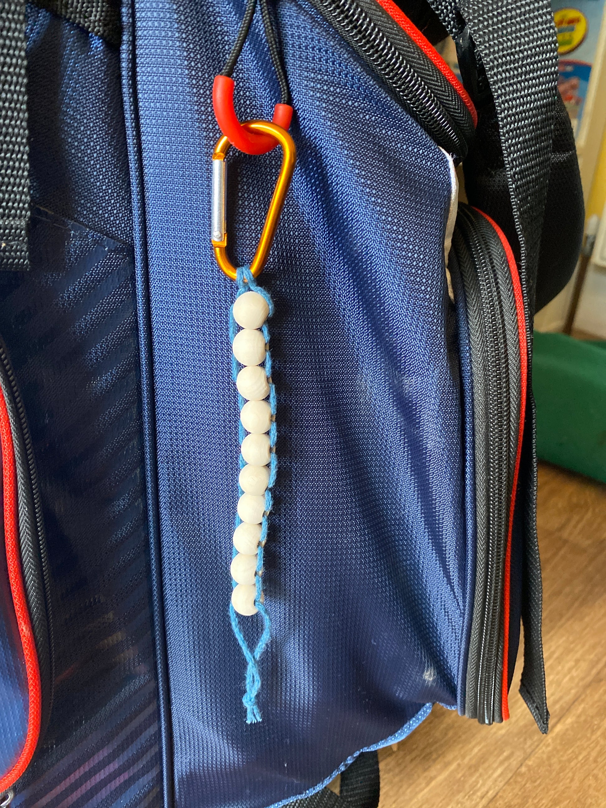 A handmade macramé golf stroke counter made with teal yarn and 10 small plain wooden beads, attached to a blue golf bag with an orange carabiner clip.