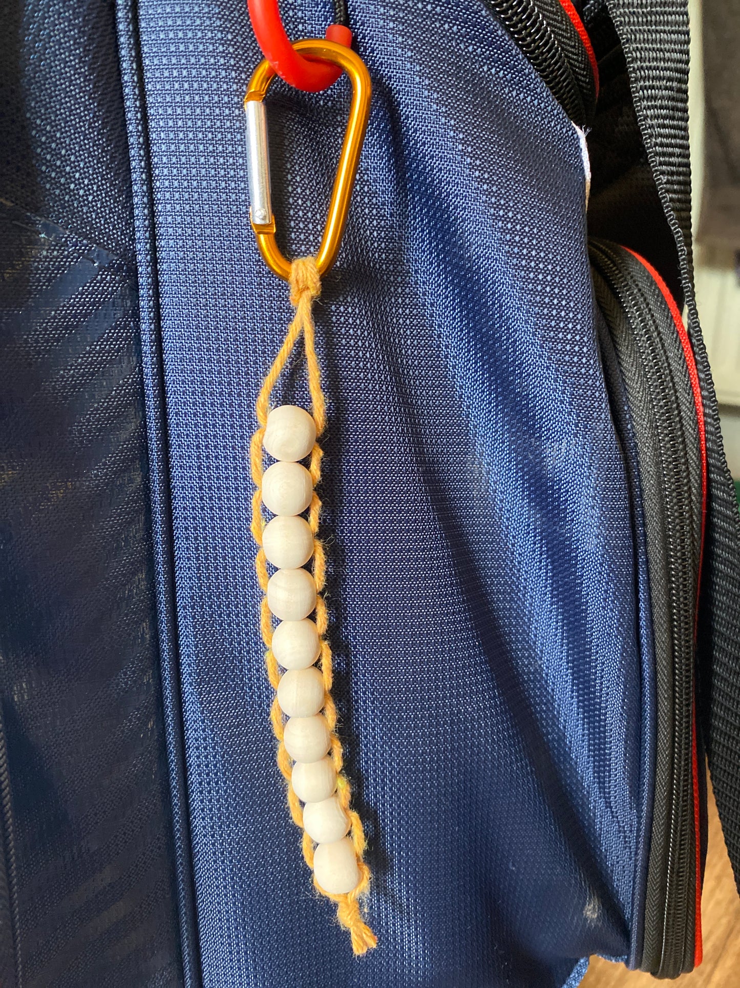 A handmade macramé golf stroke counter made with yellow yarn and 10 small plain wooden beads, attached to a blue golf bag with an orange carabiner clip.