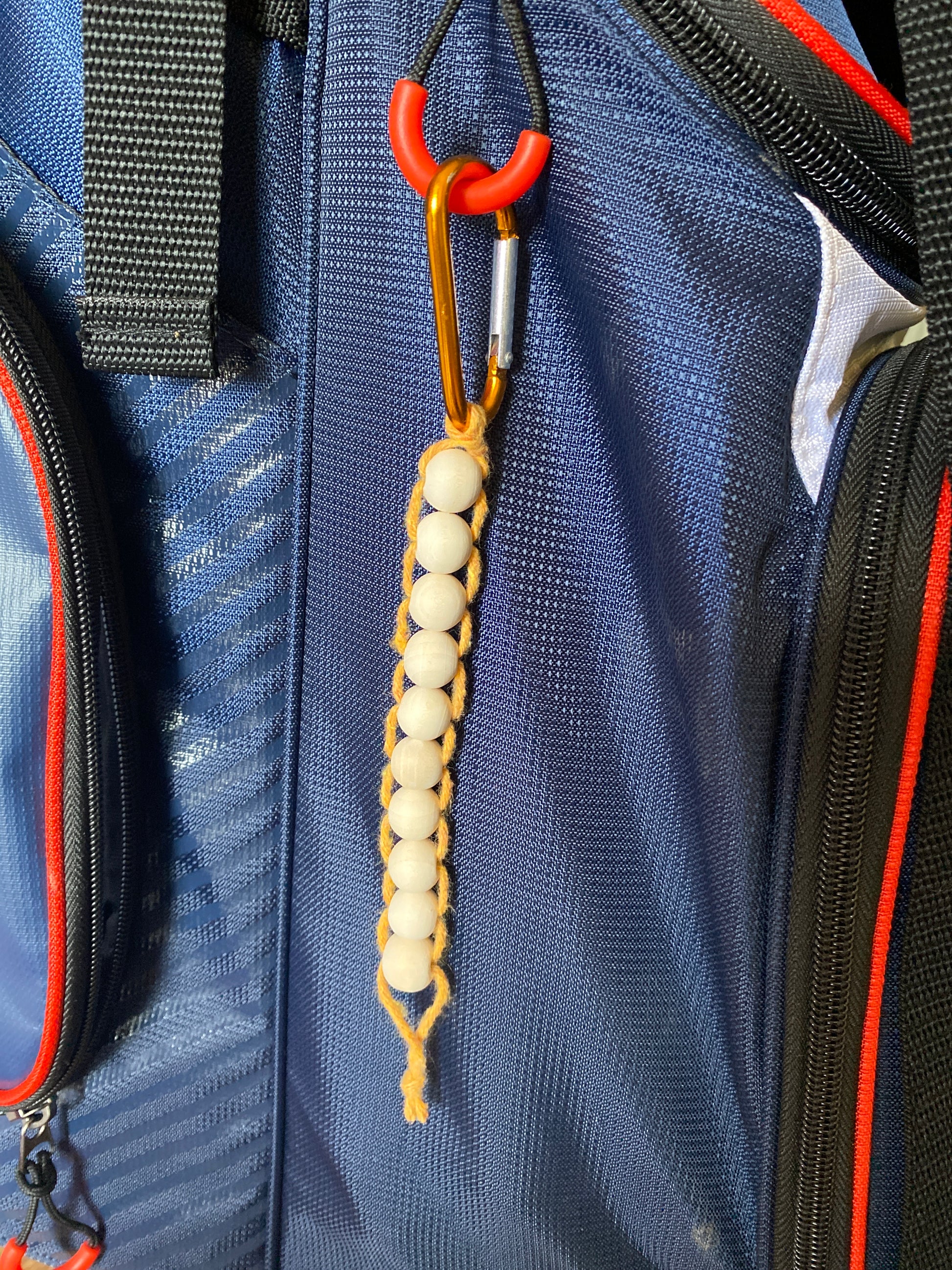 A handmade macramé golf stroke counter made with yellow yarn and 10 small plain wooden beads, attached to a blue golf bag with an orange carabiner clip.