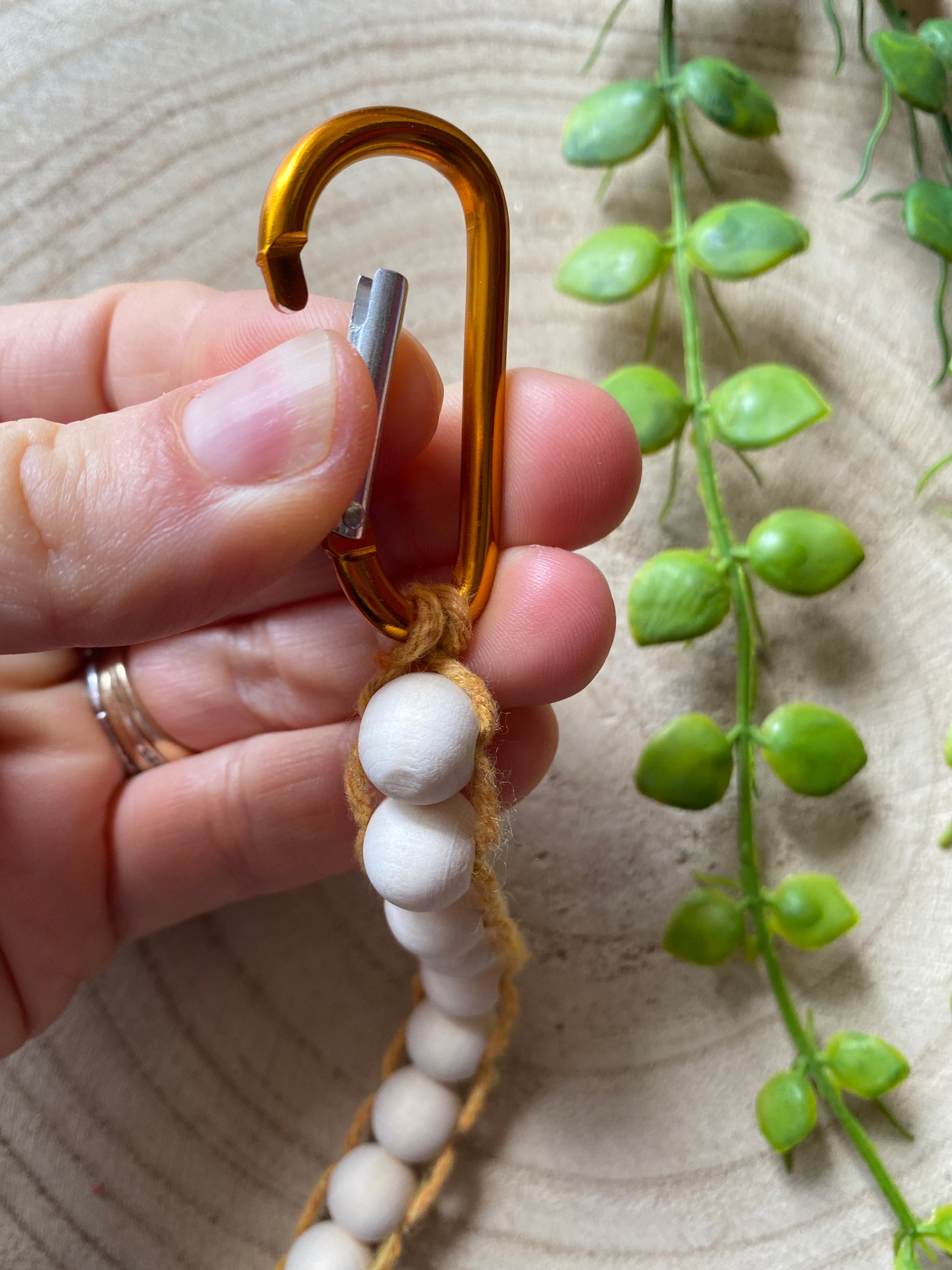 A hand demonstrating how to open an orange carabiner clip on the top of a handmade macramé golf stroke counter made with yellow yarn and small plain wooden beads.