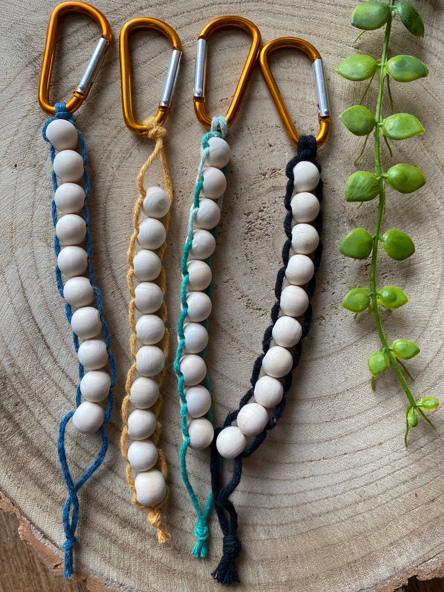 4 handmade macrame golf stroke counters lay next to each other on a wooden surface. They are made with blue, yellow, green and black yarns on orange carabiner clips, and each have 10 small wooden beads on them.