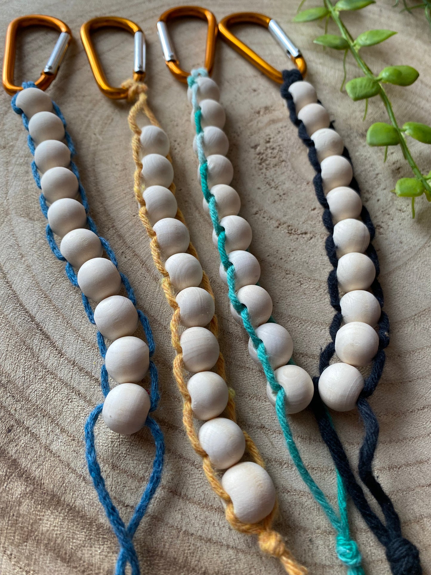 A close up of 4 handmade macrame golf stroke counters lay next to each other on a wooden surface. They are made with blue, yellow, green and black yarns on orange carabiner clips, and each have 10 small wooden beads on them.