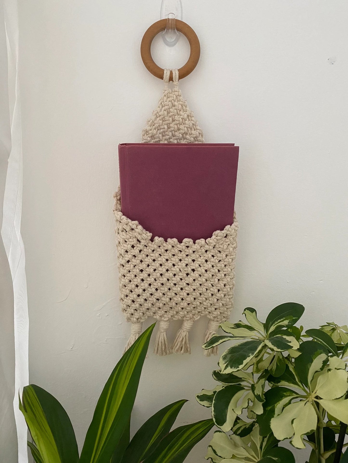 A small pink book is nestled inside a Macra-Made-With-Love Bedside Storage Pouch Book Sleeve. The cream-colored, hand-knitted wall pocket features a wooden ring at the top for hanging and tassels at the bottom. It hangs on a white wall with green plants visible in the foreground, adding recycled cotton decor charm.