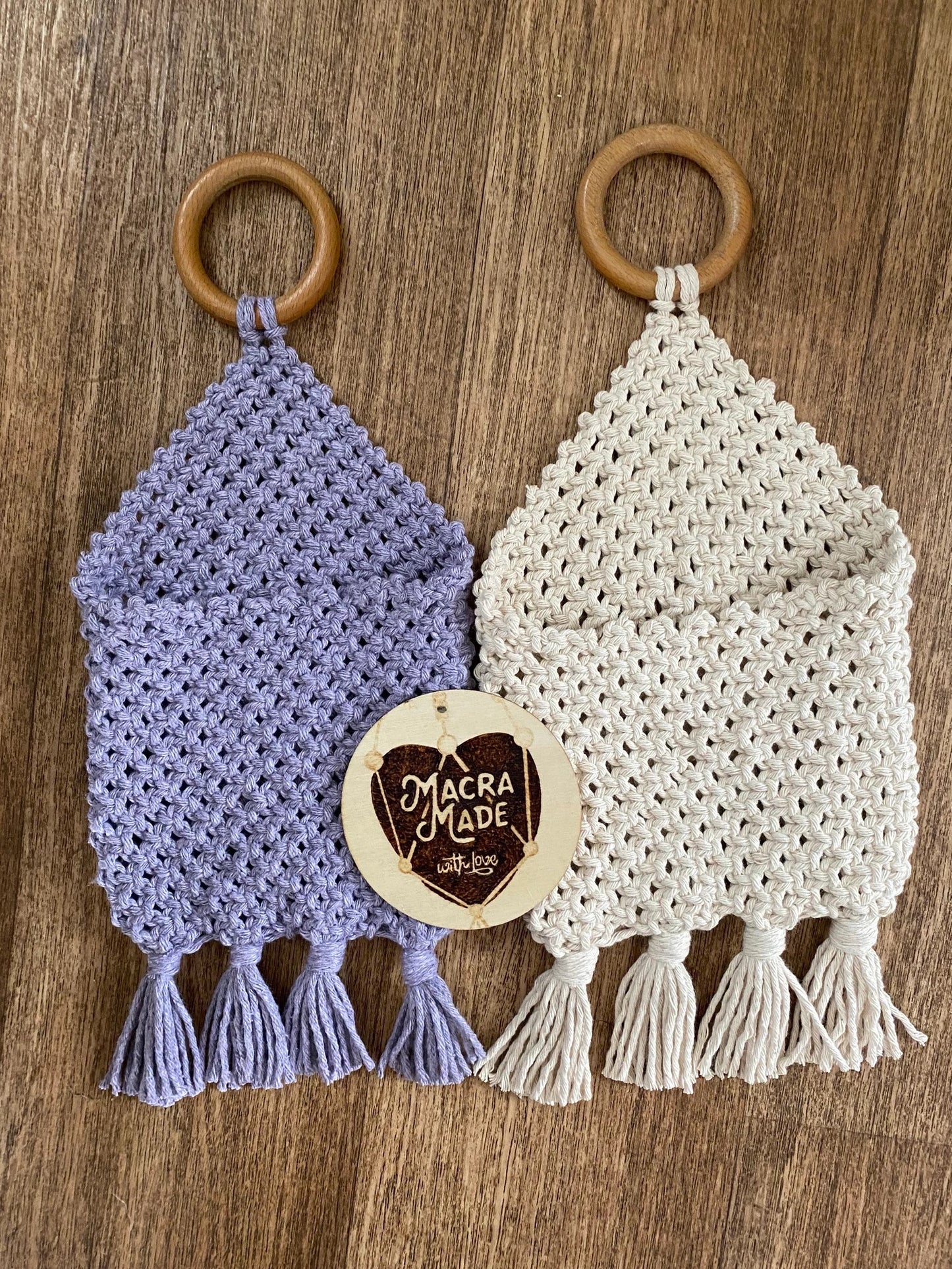 Two handmade macrame Bedside storage pouch book sleeves hang from wooden rings on a wooden surface. The left pouch is light purple, and the right pouch is off-white. Both feature intricate woven patterns and tassels at the bottom. A round "Macramade with love" tag is attached to one of the Macra-Made-With-Love Bedside storage pouch book sleeves.