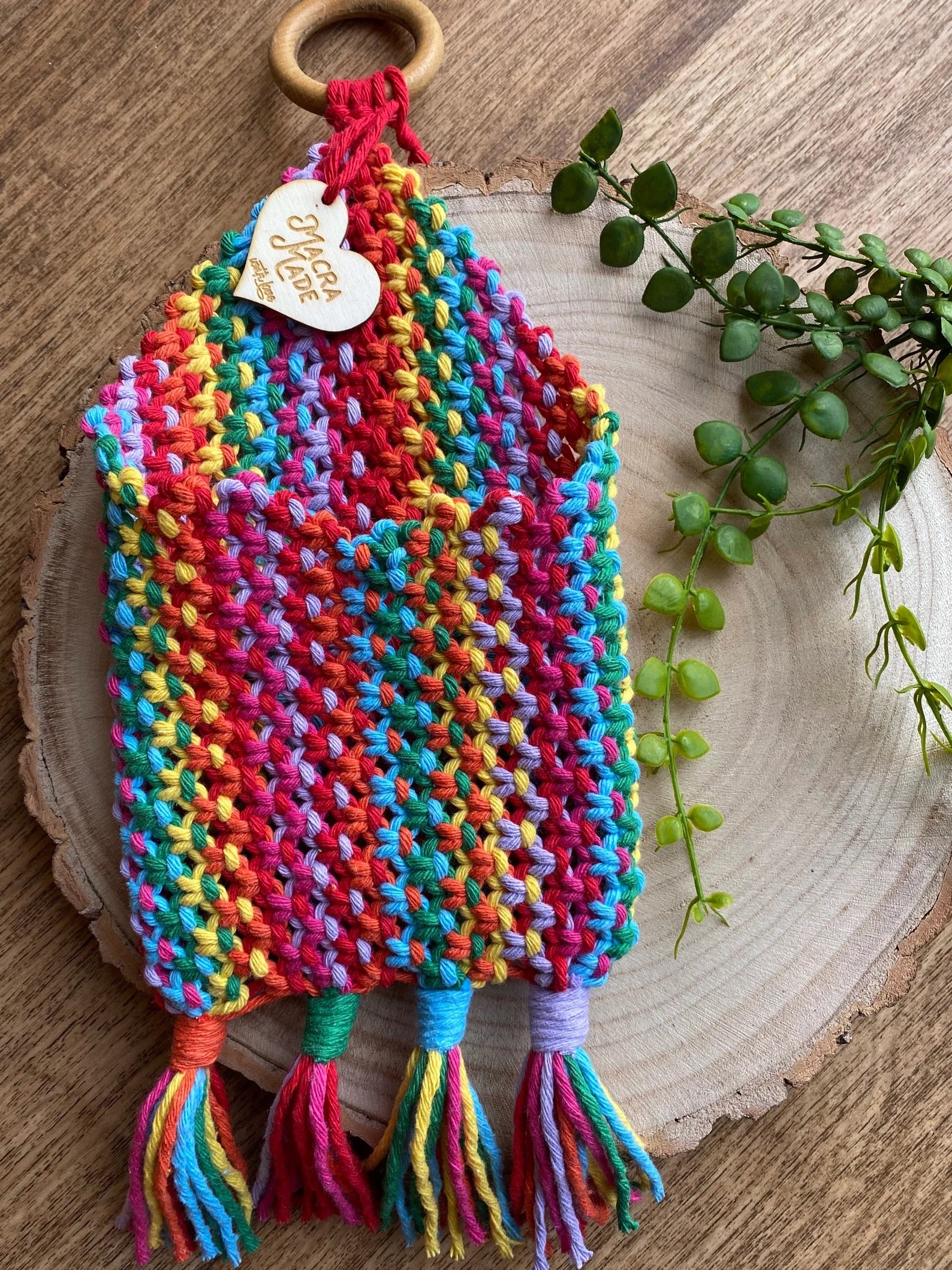 A Bedside storage pouch book sleeve by Macra-Made-With-Love crafted from recycled cotton decor features a wooden ring at the top and a small heart-shaped tag reading "Handmade with Love." This vibrant piece boasts rainbow colors and tassels at the bottom, displayed on a wooden surface with a small green plant nearby.
