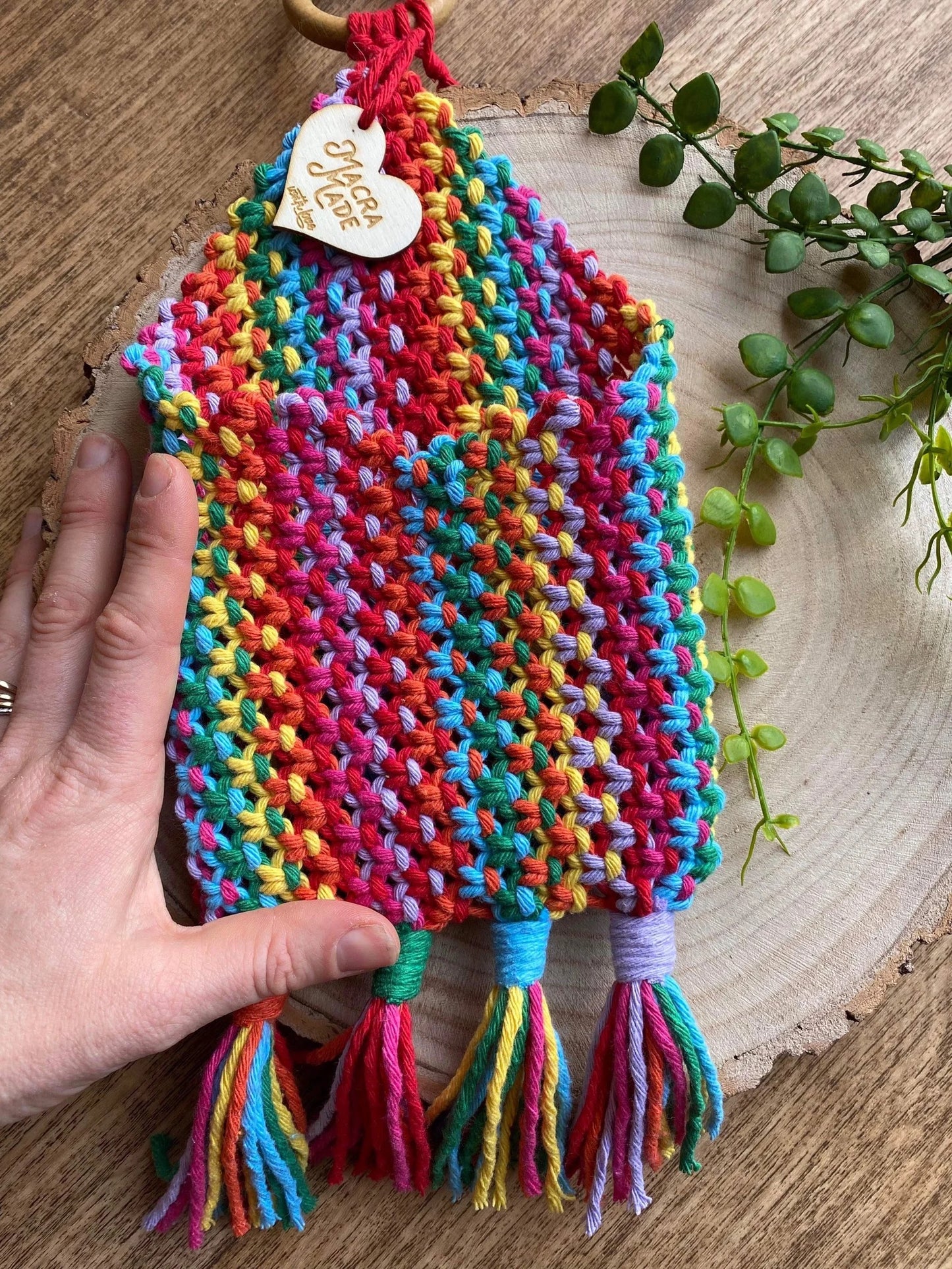 A hand demonstrates a colorful, handmade Bedside storage pouch book sleeve adorned with multiple vibrant hues of yarn and tassels at the bottom. The pouch, crafted from recycled cotton decor by Macra-Made-With-Love, is displayed on a wooden surface next to a leafy green plant and has a wooden tag attached with an inscription.