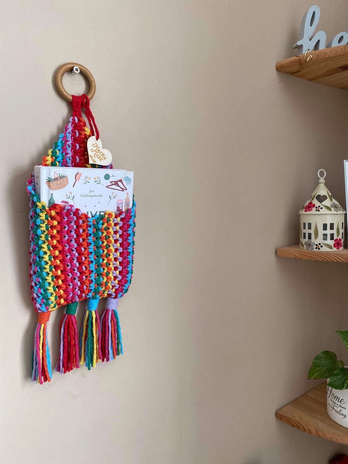 A colorful Bedside storage pouch book sleeve by Macra-Made-With-Love, made from recycled cotton decor, is mounted on a beige wall by a wooden ring. It holds a folded magazine and features multicolored tassels at the bottom. Wooden shelves on the right display a decorative house lantern, a small plant, and other decor.