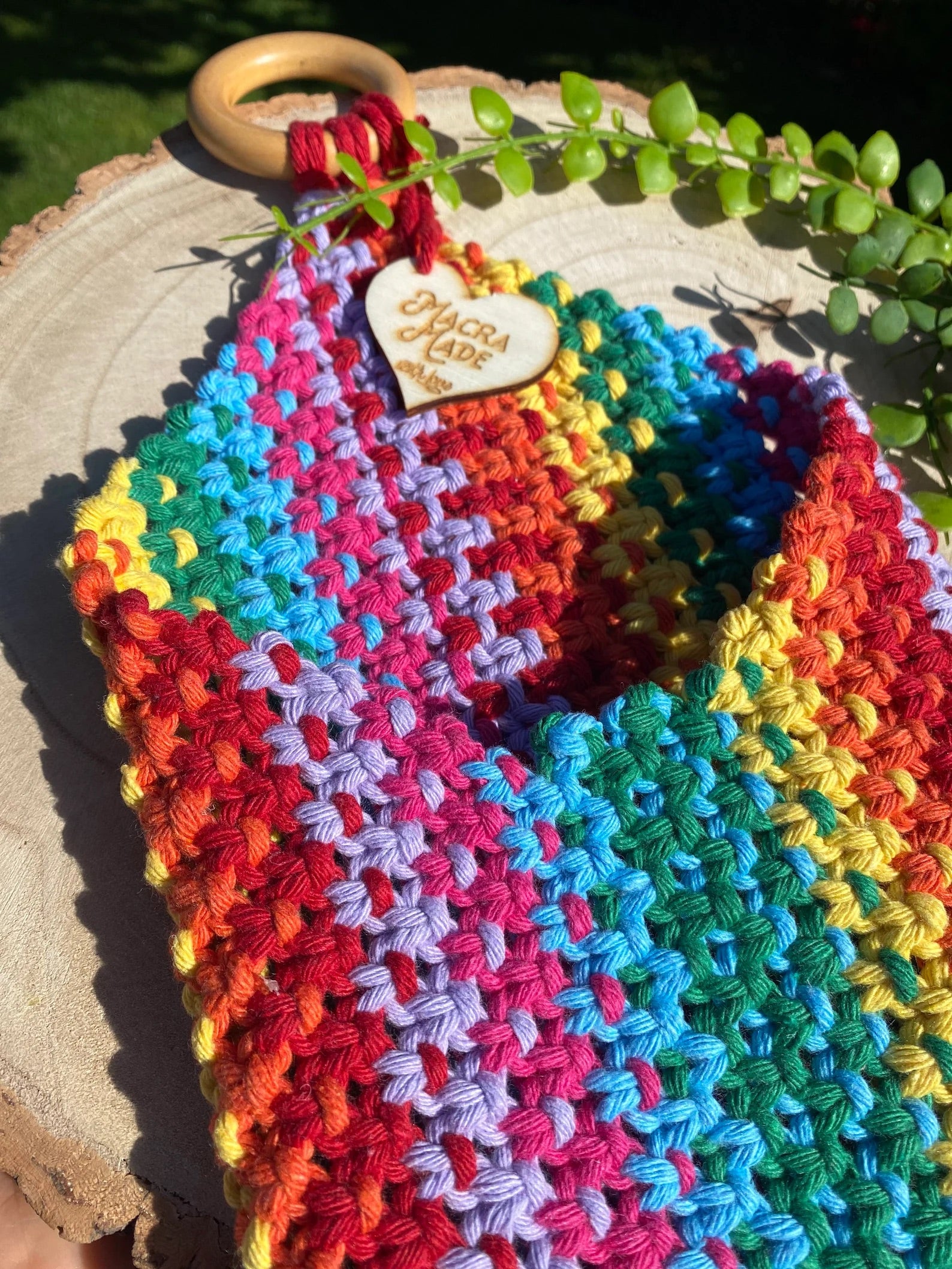 Colorful crocheted fabric in a rainbow pattern, resting on a wooden surface. A wooden ring is attached to one end of the fabric with a charm that reads "Handmade with Love." The Bedside Storage Pouch Book Sleeve by Macra-Made-With-Love doubles as an eco-friendly bedroom storage solution. A green succulent plant is visible in the background, adding a natural touch.
