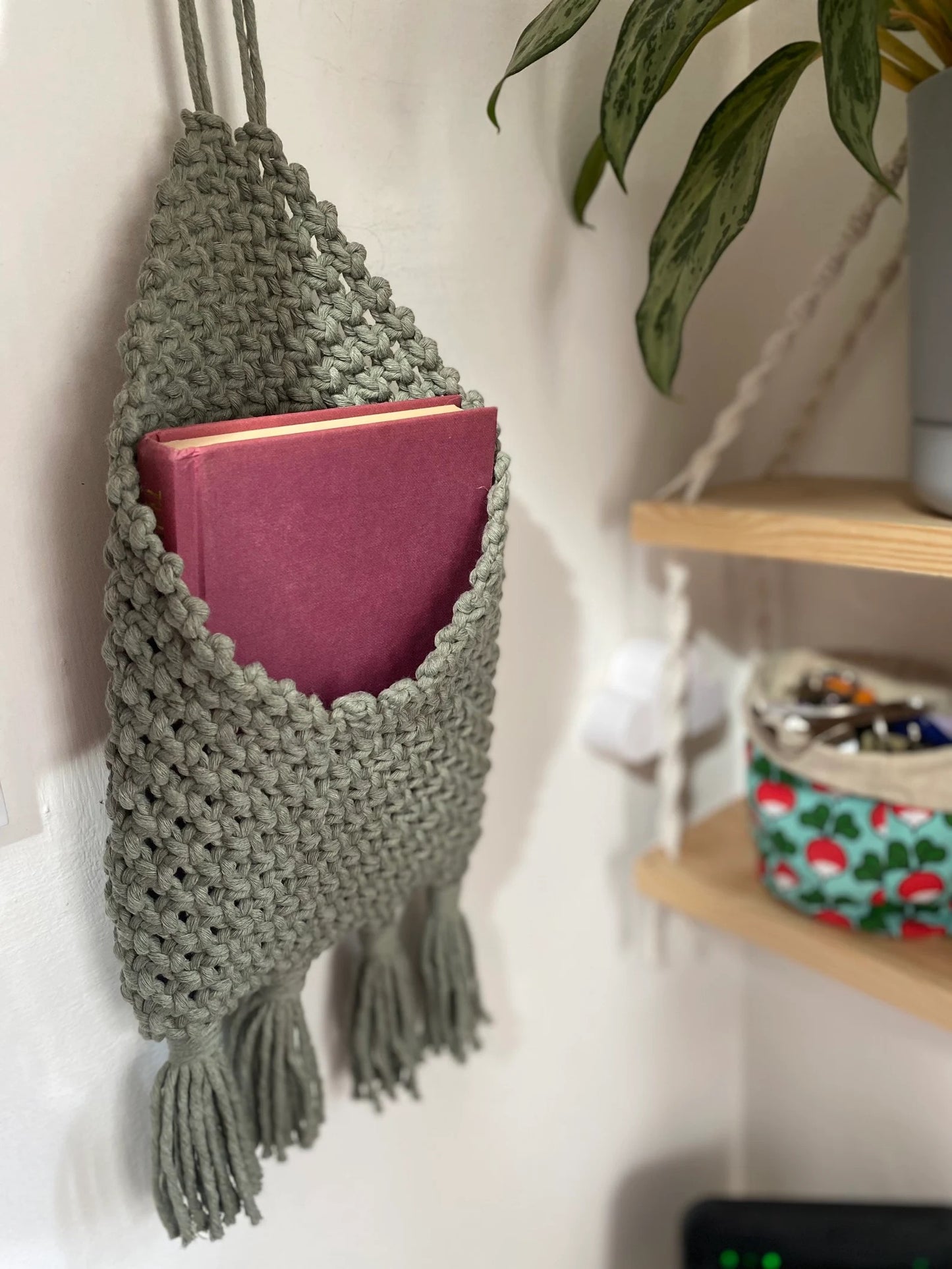 A handmade macrame storage pouch in light green hangs on the wall, holding a closed maroon book. Below it is a wooden shelf with decorative items, including a small bowl containing various items and a plant. Part of a larger plant is visible to the upper right, creating eco-friendly bedroom storage with the Macra-Made-With-Love Bedside Storage Pouch Book Sleeve.