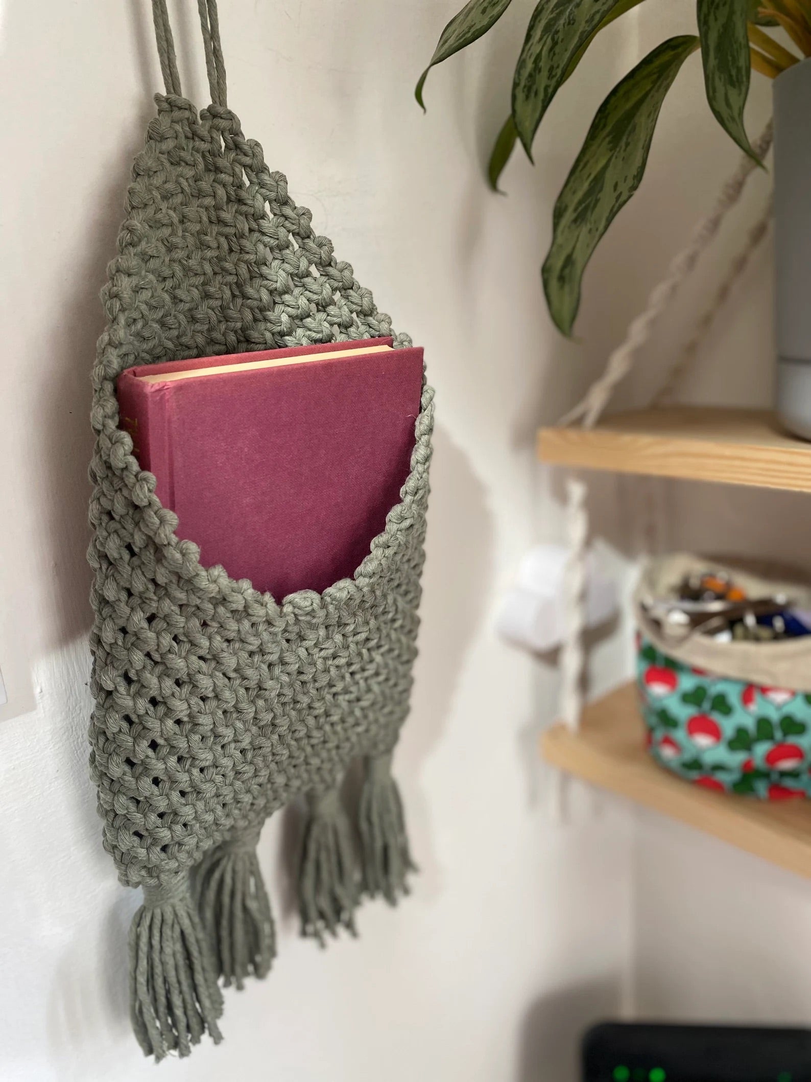 A handmade macrame storage pouch in light green hangs on the wall, holding a closed maroon book. Below it is a wooden shelf with decorative items, including a small bowl containing various items and a plant. Part of a larger plant is visible to the upper right, creating eco-friendly bedroom storage with the Macra-Made-With-Love Bedside Storage Pouch Book Sleeve.