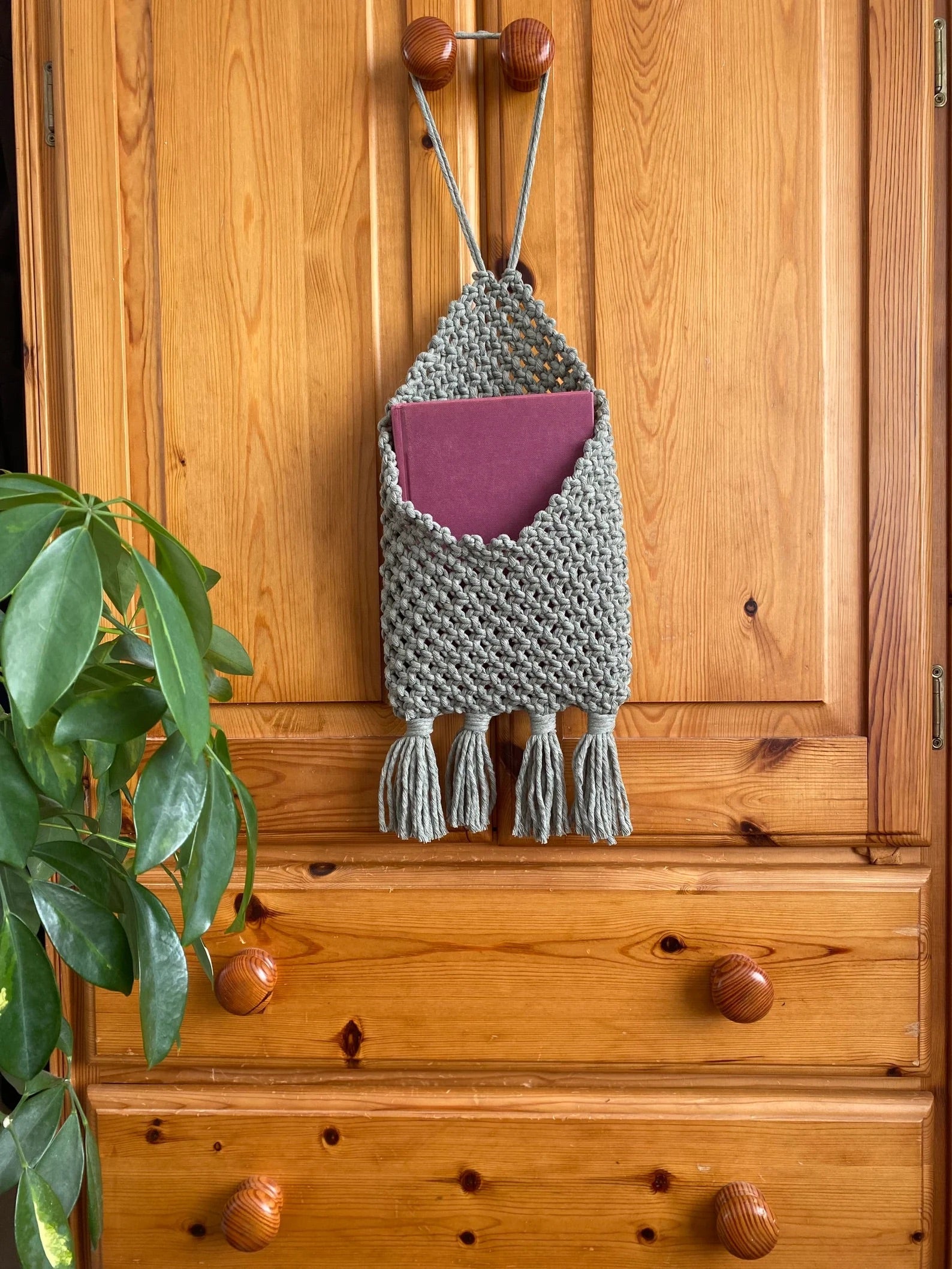 A wooden dresser with two round-knob handles features a gray handmade macrame storage pouch with fringes, holding a maroon book. A green leafy plant is partially visible on the left side of the image, adding a touch of nature to this eco-friendly bedroom storage solution. This Bedside storage pouch book sleeve by Macra-Made-With-Love combines style and functionality seamlessly.