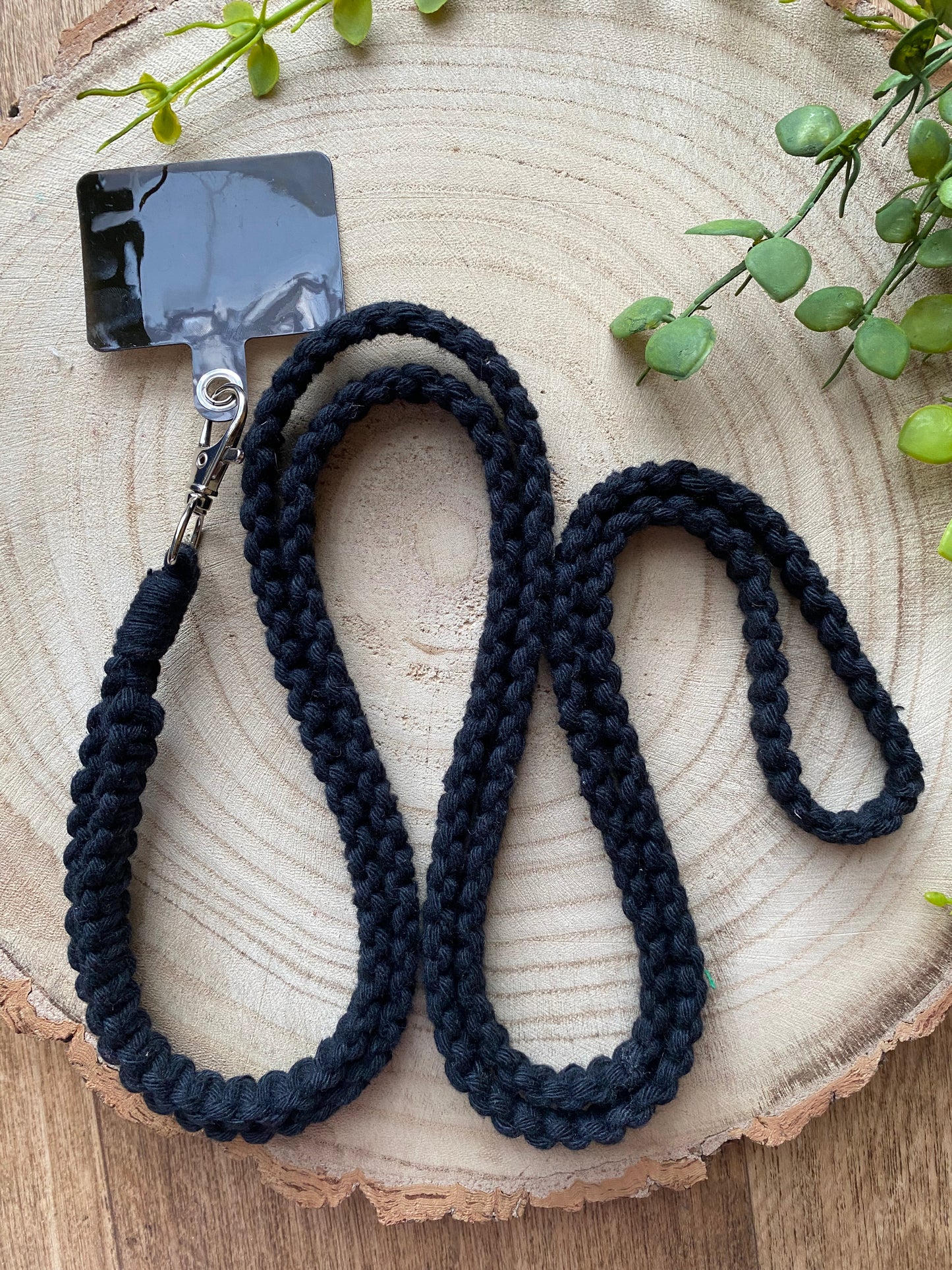 A black braided lanyard with a metal clasp holding a rectangular black badge holder is coiled on a wooden surface, resembling the intricate beauty of **a Macra-Made-With-Love recycled cotton crossbody phone strap**. Green plants are placed around the edge of the wooden surface for decoration, enhancing its eco-friendly charm.